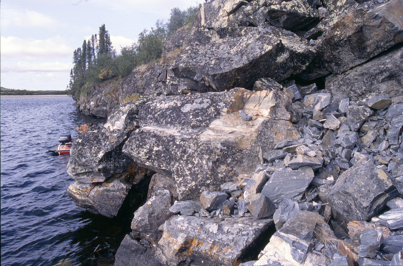 A cliff outcrop of light and dark rocks with water on the left (the stern of a small red boat can just been seen). Cliff is around 10 metres In height with a jumble of large lichen covered rounded boulders on the left and a patch of small angular clean looking rocks on the right. This is the location of the reputed explosion to collect rocks.