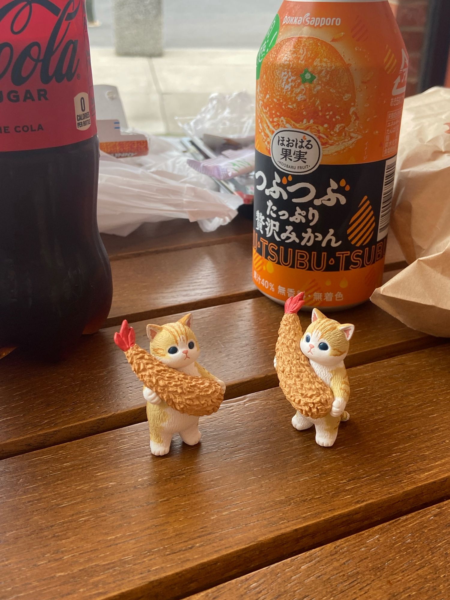 two small cat figurines holding tempura shrimp on a wooden table in front of a bottle of tsubu-tsubu orange drink and coke zero