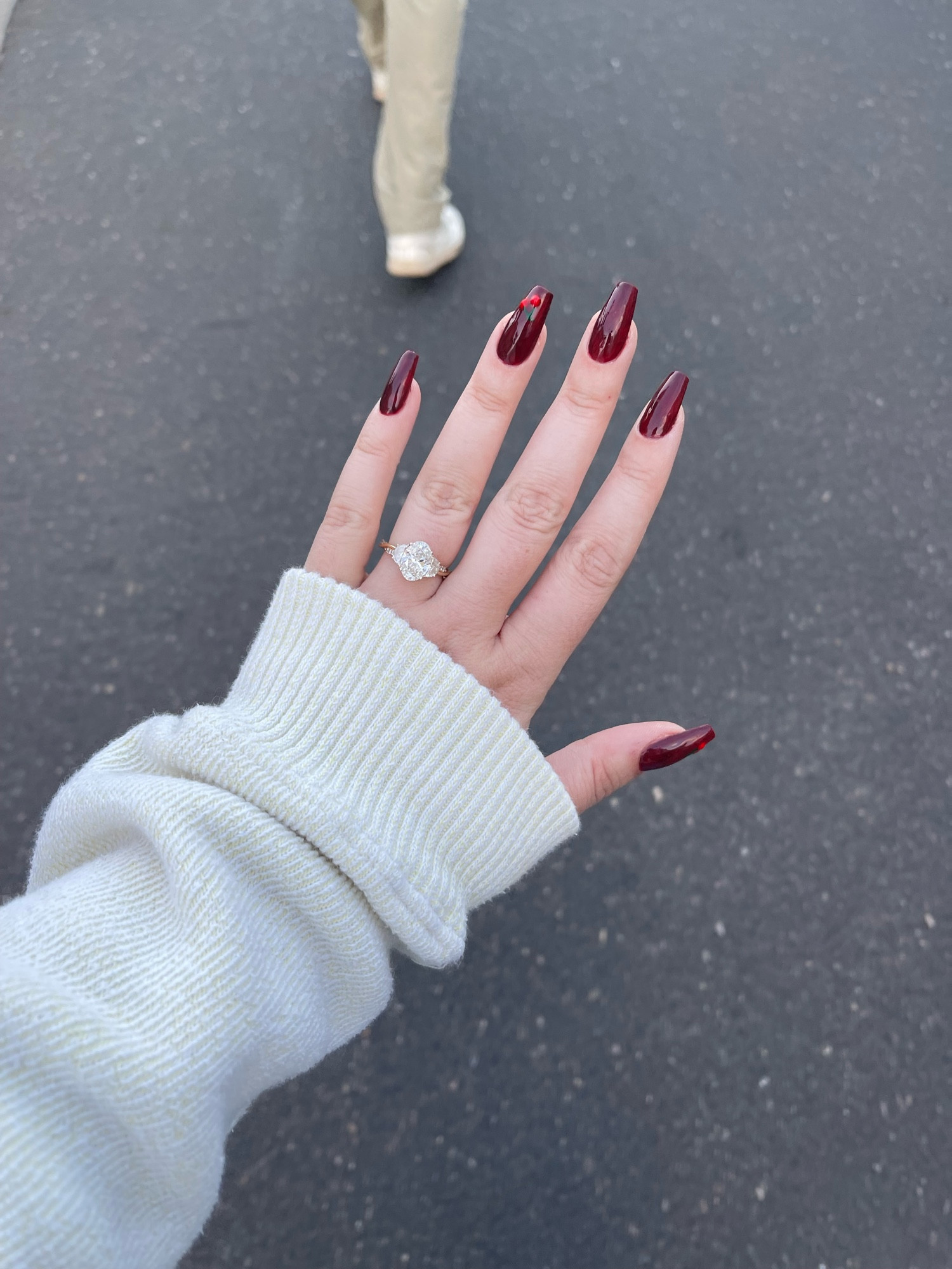 A left hand with a cream colored sleeve. Background is black pavement. Nails are a medium coffin shape with a reddish – brown color “cherry Coke nails” with a cherry design on the ring finger and thumb.