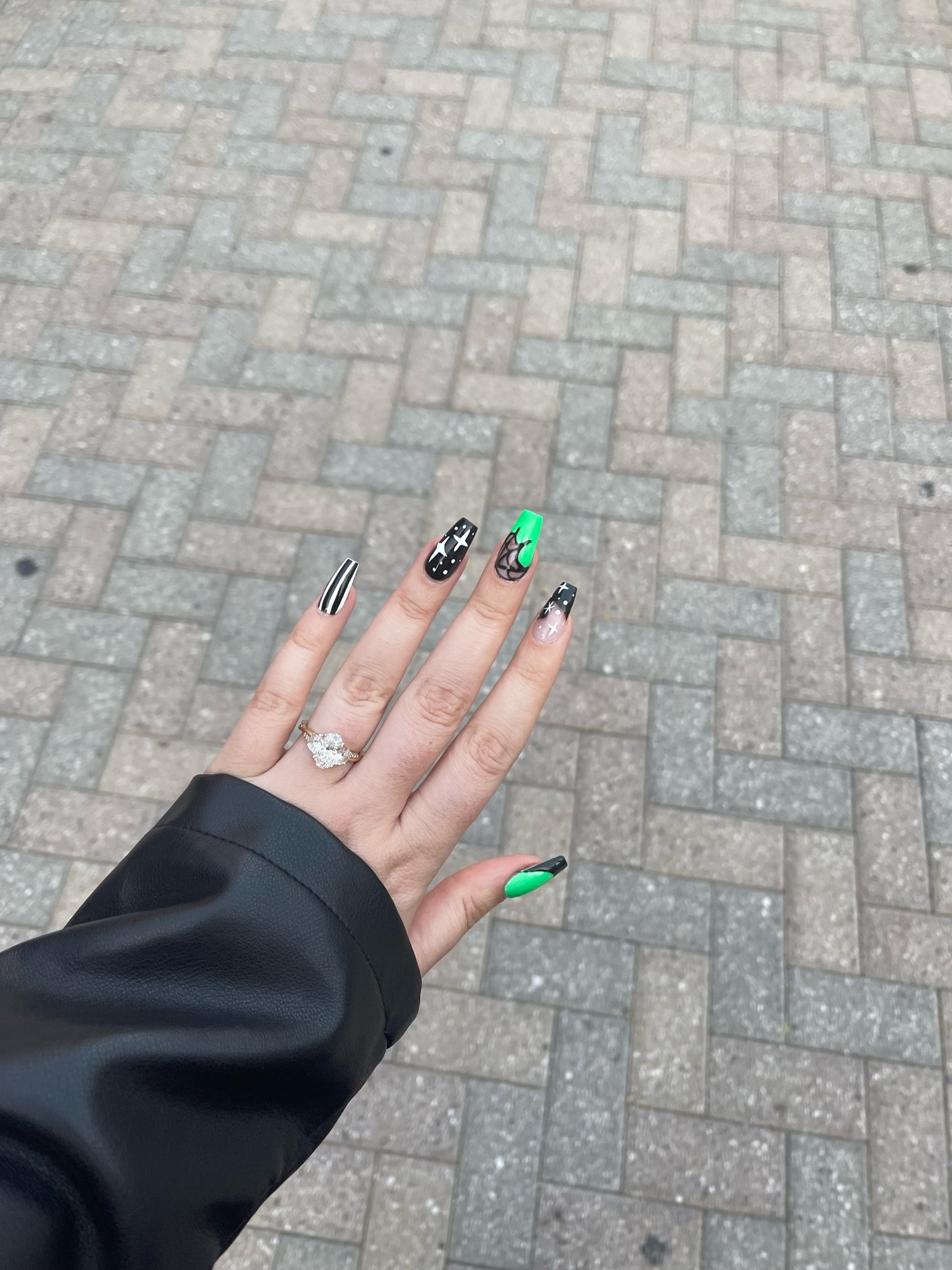 A left hand with a black pleather sleeve. Background is gray, brick, pavement nails are medium coffin. Nail designs feature black, white and green Beetlejuice theme.