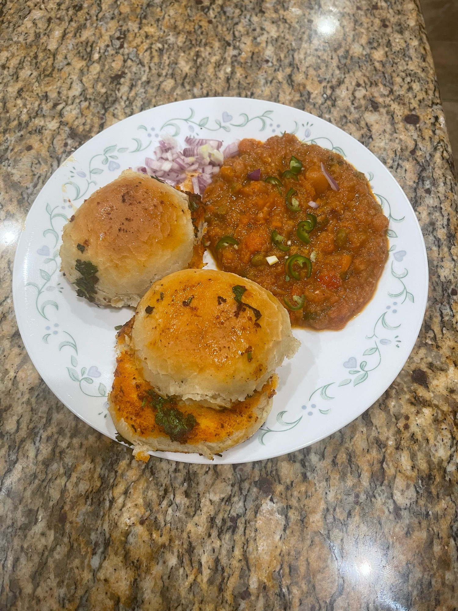 two rolls made of white flour, an Indian recipe called “pav” - pronounced “puv”
on a white plate with a healthy scoop of bhaji, a mumbai street-style curry. topped with green pepper and cilantro. 