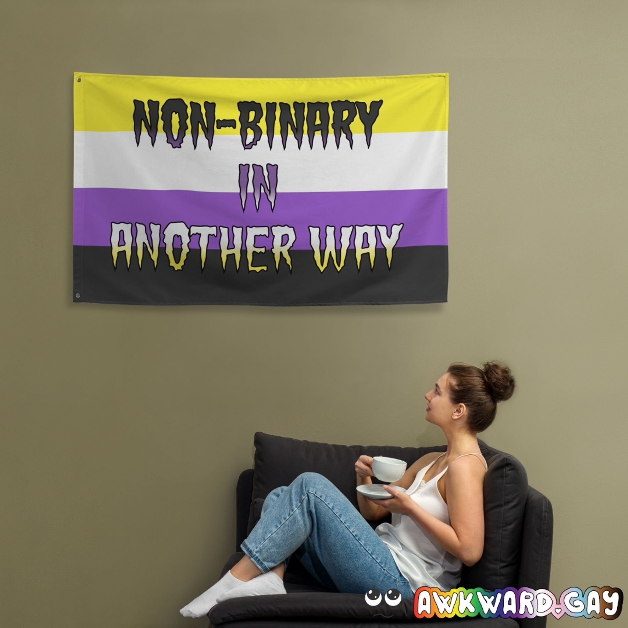 Mockup of a flag hanging on a wall, with someone sitting below looking up at it. The flag features the non-binary pride flag and the words "non-binary in another way"