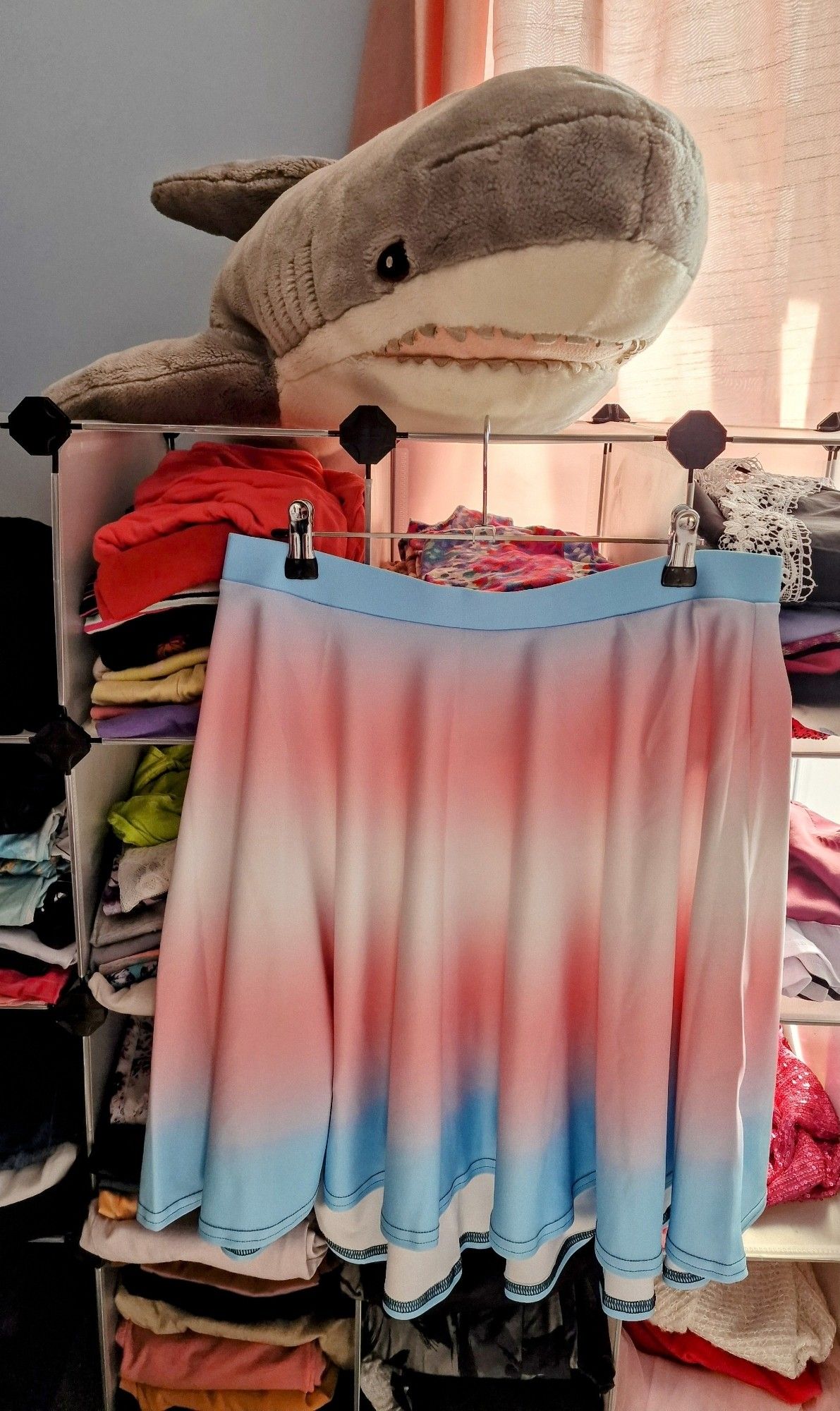 A skater skirt with a trans pride flag gradient print, hanging on a cube storage rack full of clothes, with a grey Blåhaj sitting on top