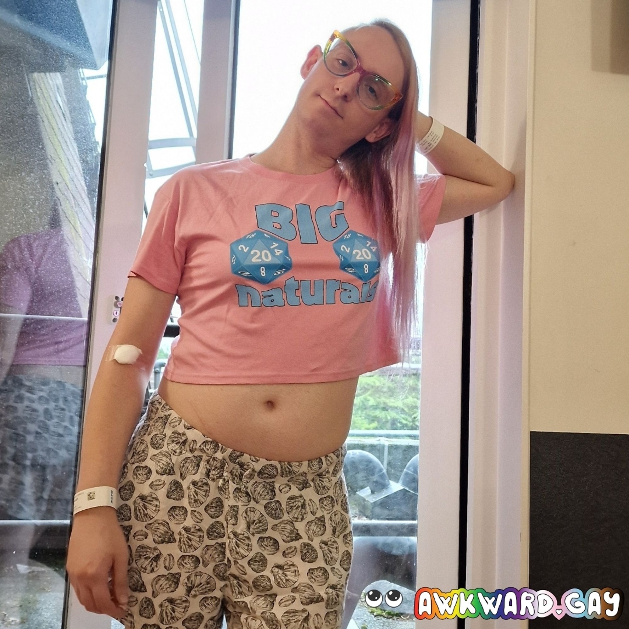 Me stood in front of slightly open glass doors in a hospital room wearing a pink crop top with a blue graphic (detail in next pic). I'm leaning against the door frame with my left elbow with my head resting against my hand. I have a cotton wool ball taped to my right inner elbow where I had blood drawn, and an id bracelet on each wrist