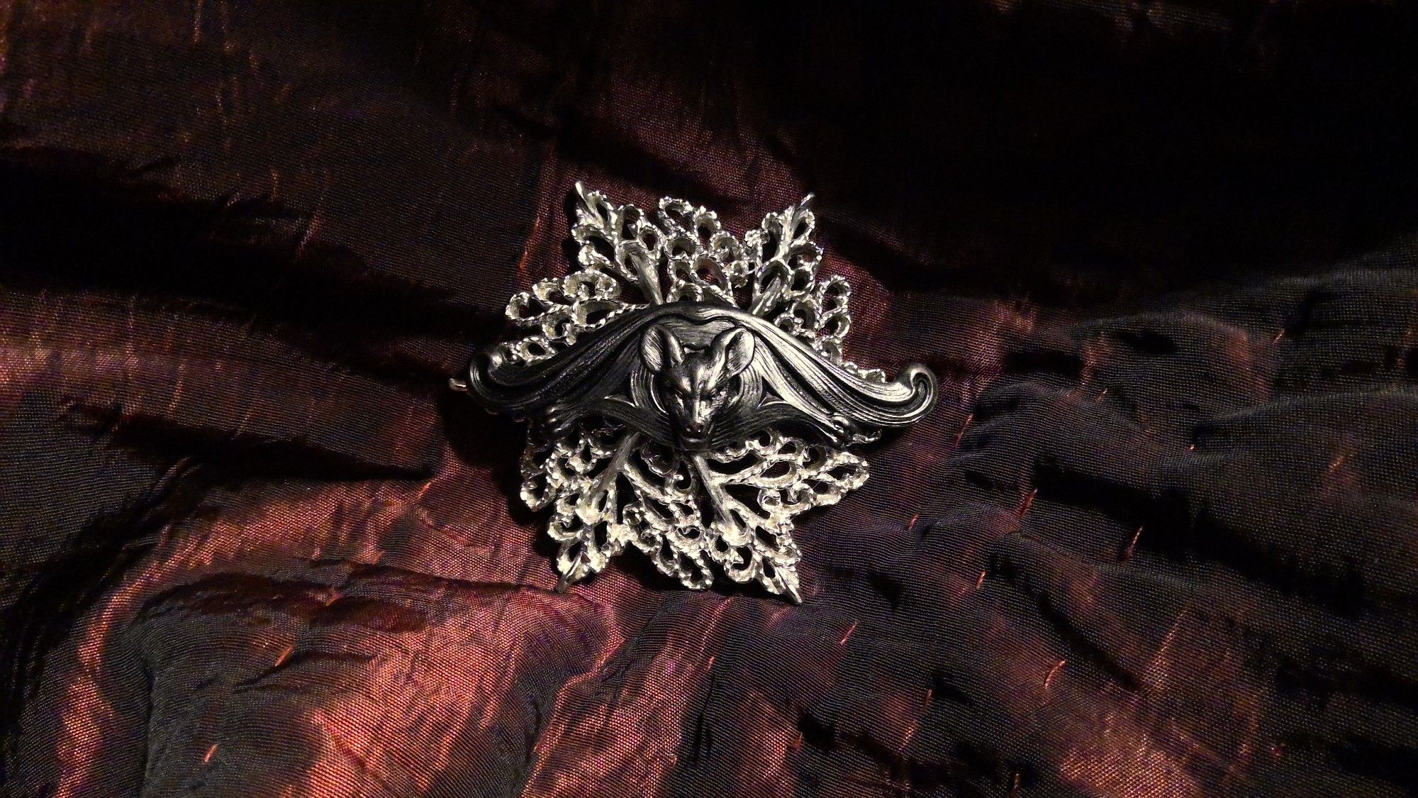 A silver-tone leafy, fractal-like-patterned brooch, like a feathery starburst, with a blackened brass Art Deco bat at the center of the brooch. Approx 2 by 2 inches