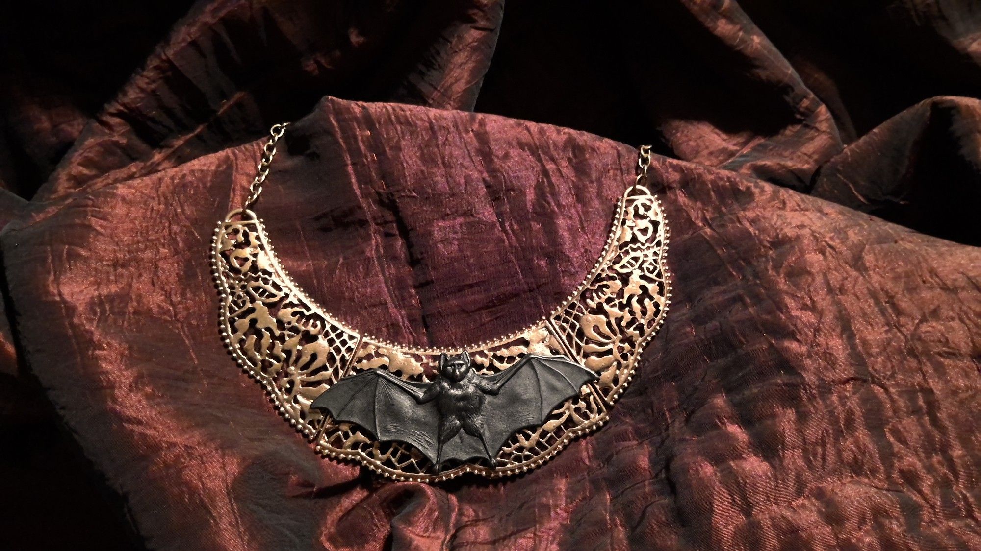 A golden-tone brass cut-lace-metal bib necklace with a large, bold, blackened brass bat at the center. Approx 3 inches tall, 6 inches wide with the cut lace, approx 18 inches of chain / give. Adjustable.