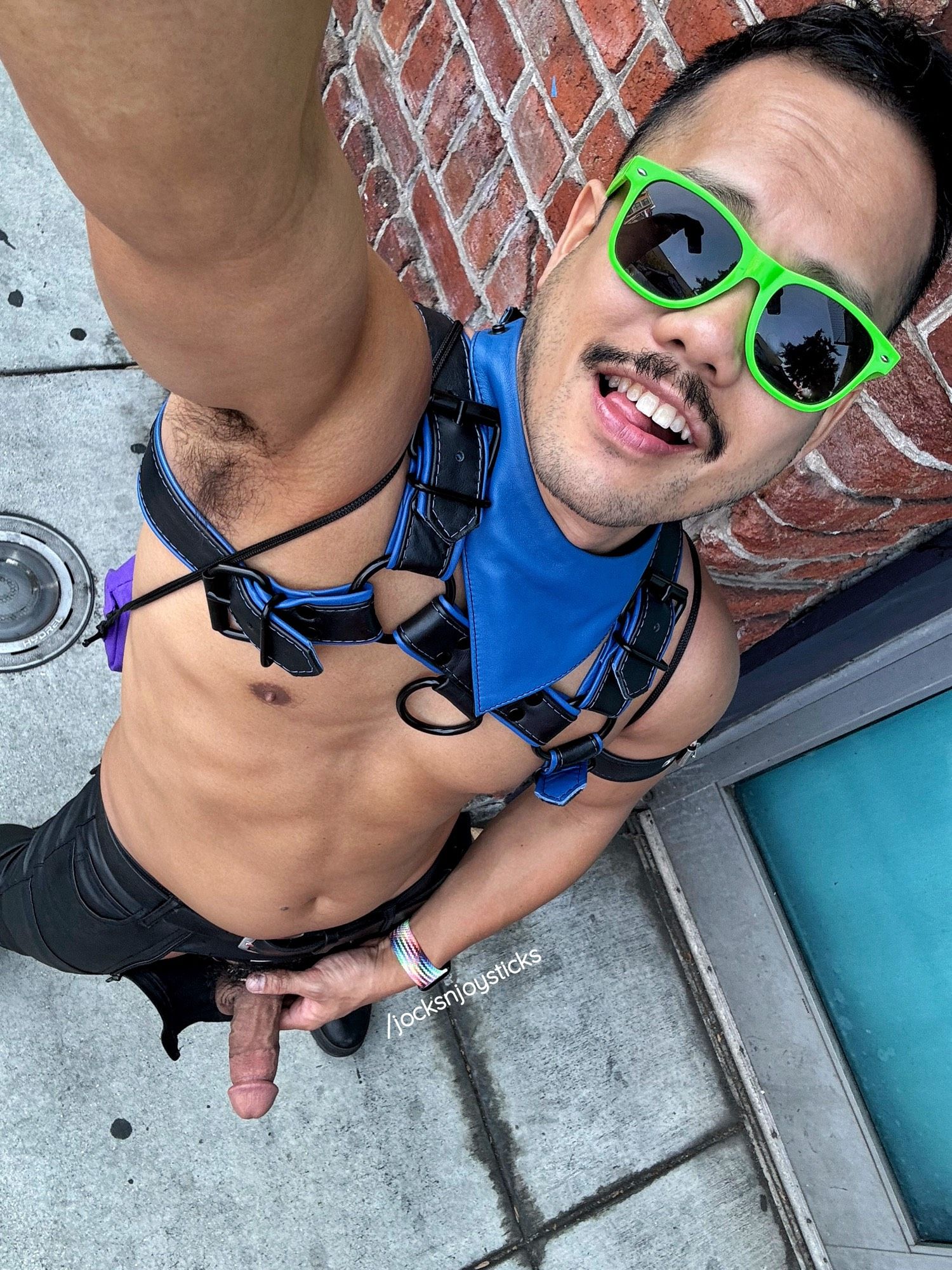 I’m outside of a brick building, taking a selfie with my camera pointing downward from overhead. I’m shirtless with a black & blue leather harness, blue leather bandana, lime green sunglasses, and shiny, black pants. The front flap of my pants are zipped open and I’m holding my hard cock in one hand. I’m grinning to the camera while biting the tip of my tongue