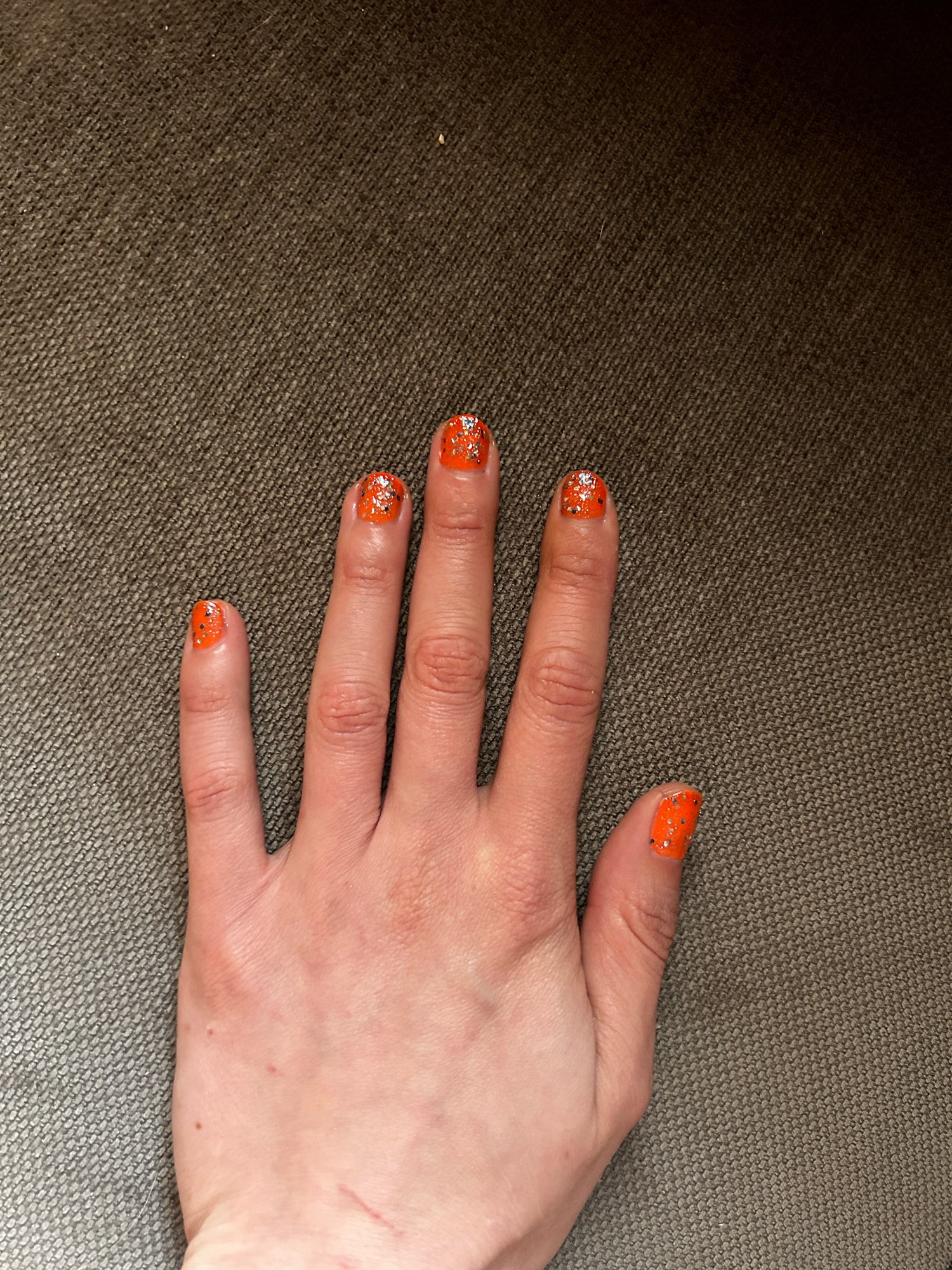 Meine linke Hand auf grauem stoffuntergrund. Meine Nägel sind neonorange lackiert mit einem überlack mit größeren und kleineren Glitzerpartikeln