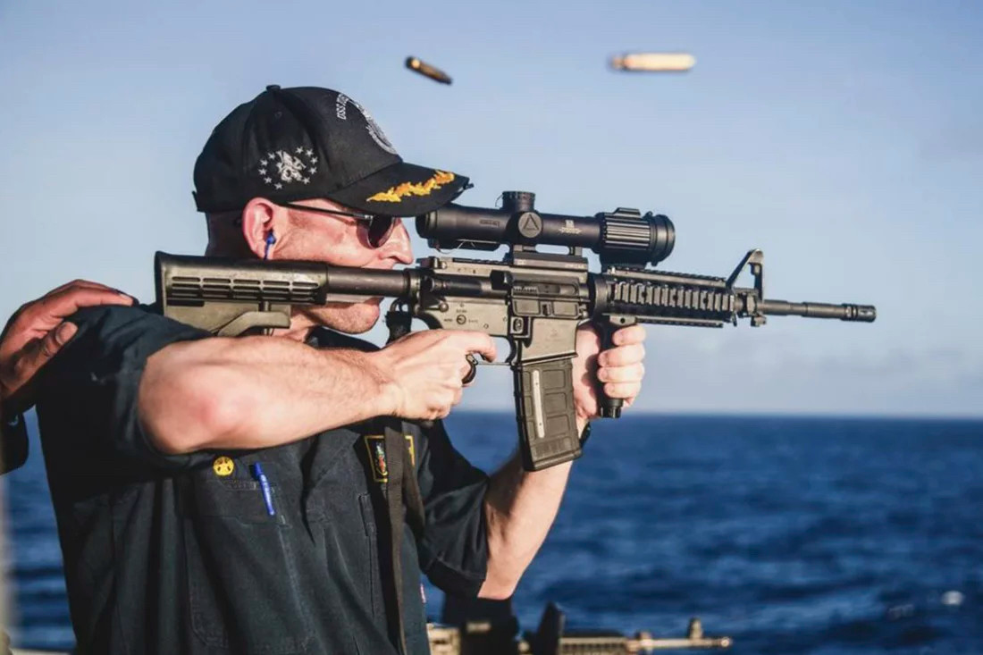 Commander Yaste firing an AR-type rifle with the scope installed backwards, such that it makes the target appear farther away 

Ejected cartridges are visible in midair, and the ocean is in soft focus behind him 