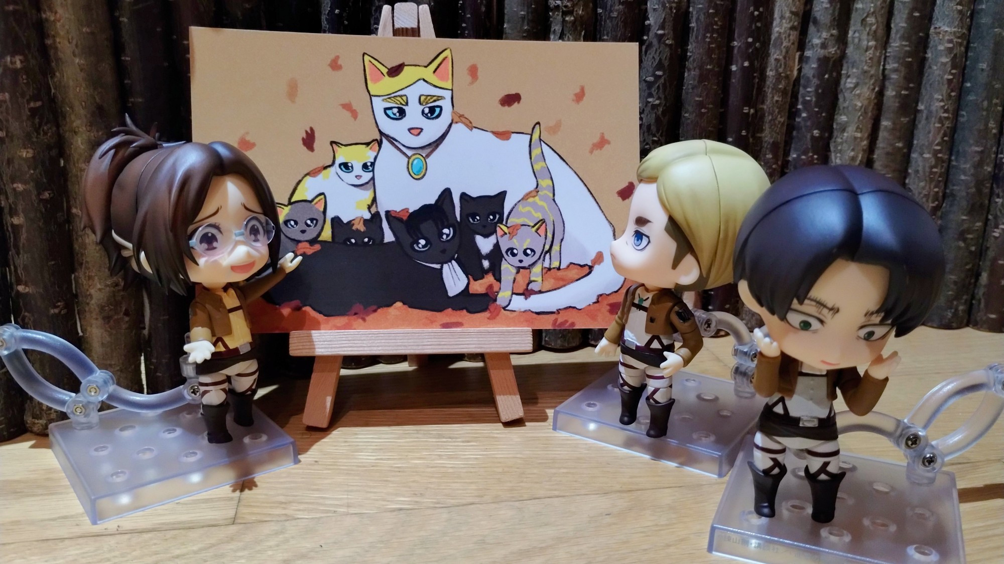 Nendoroids of Erwin Smith, Levi and Hange Zoe stand around a painting of a family of cats. Hange is smiling and gesturing at the painting as if pointing out to Erwin and Levi that the adult cats in the image resemble them. Erwin is taking another look at the painting. Levi is looking away with his head in his hands and an embarrassed expression.