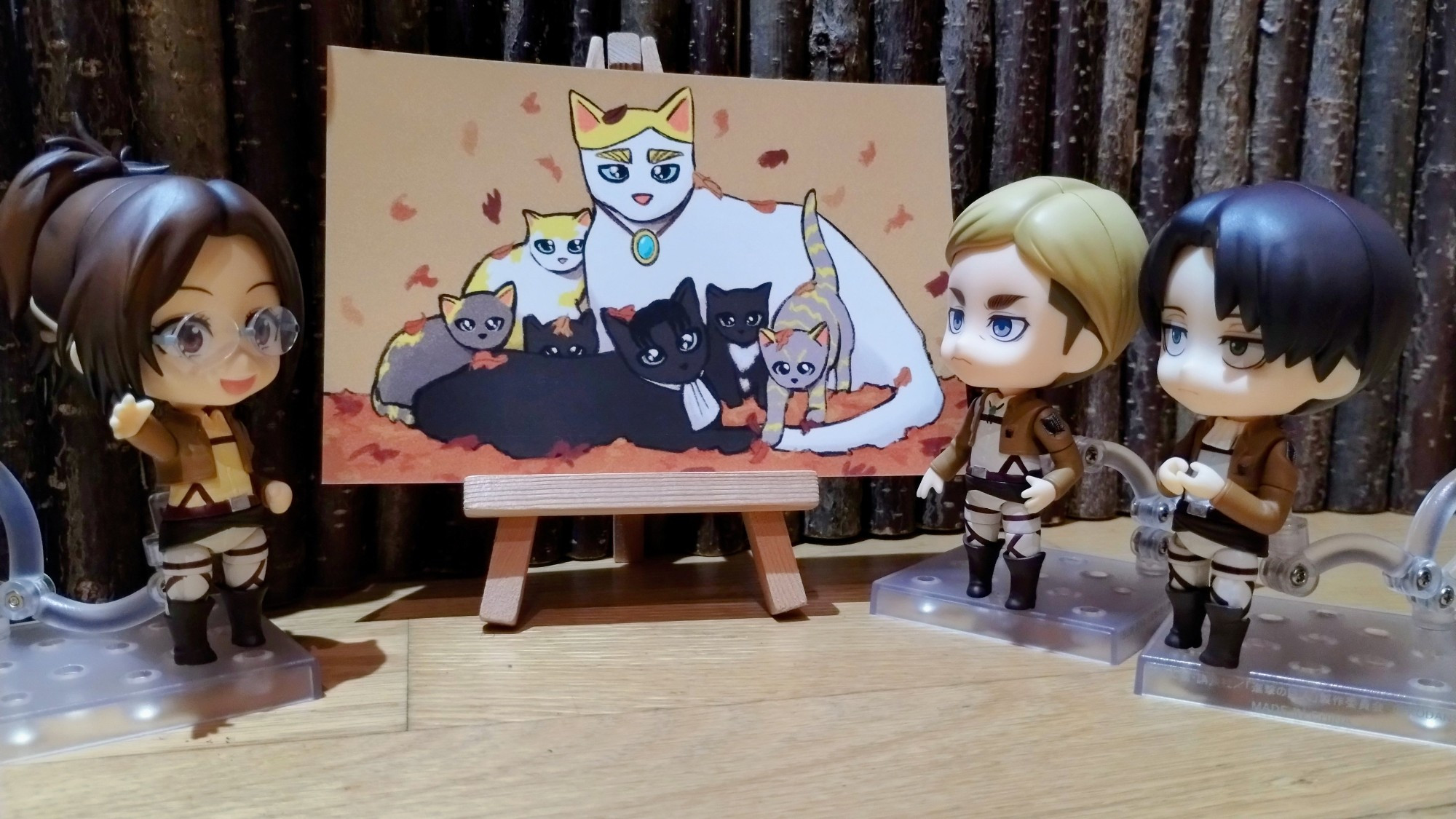 Nendoroids of Erwin Smith, Levi and Hange Zoe stand around a painting of a family of cats. Hange is smiling and waving to Erwin and Levi as though they have just arrived.