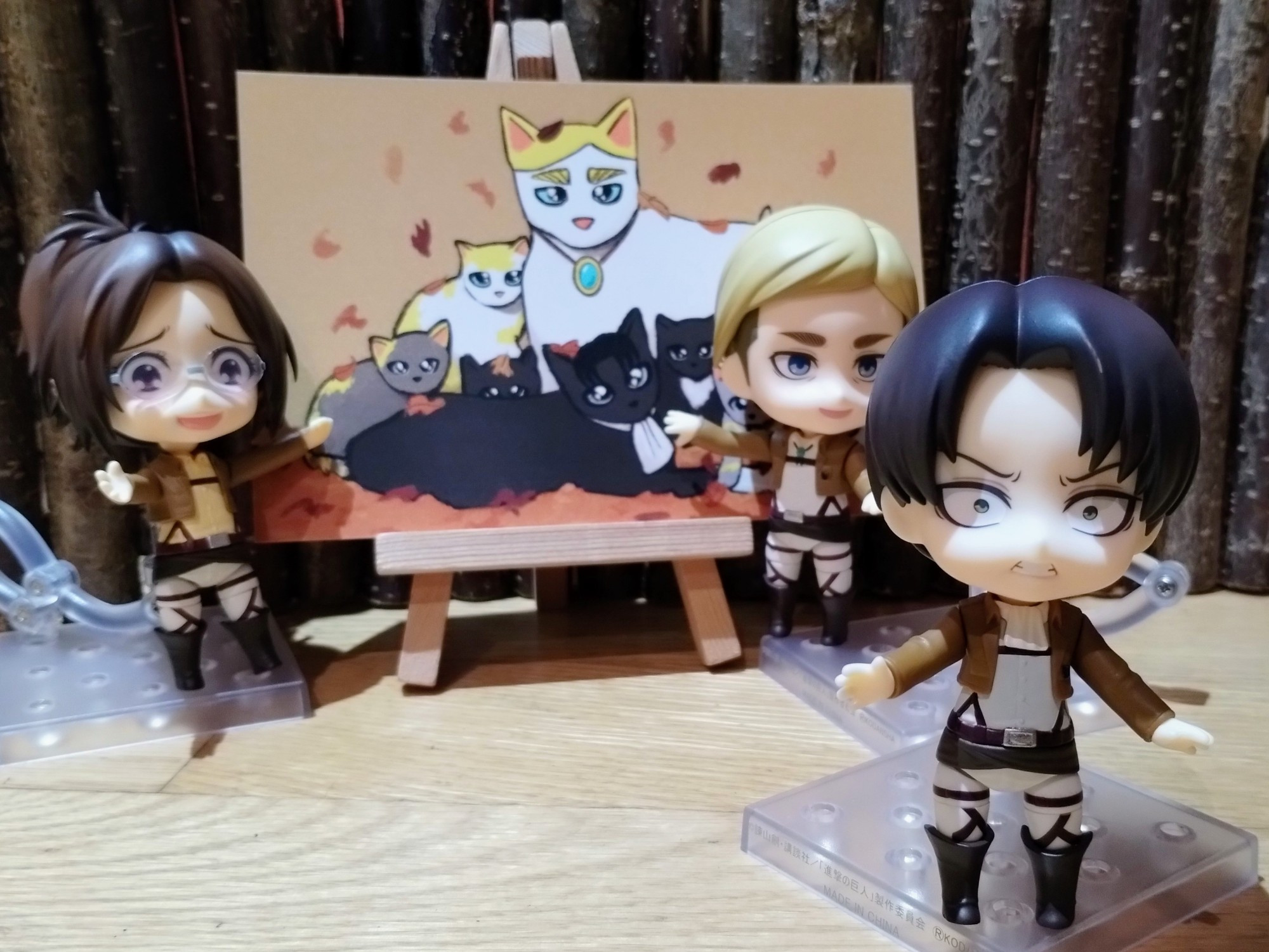 Nendoroids of Erwin Smith, Levi and Hange Zoe stand around a painting of a family of cats. Both Hange and Erwin are smiling at Levi while gesturing at the painting. Levi is walking away with his arms outstretched and a look of shock on his face.