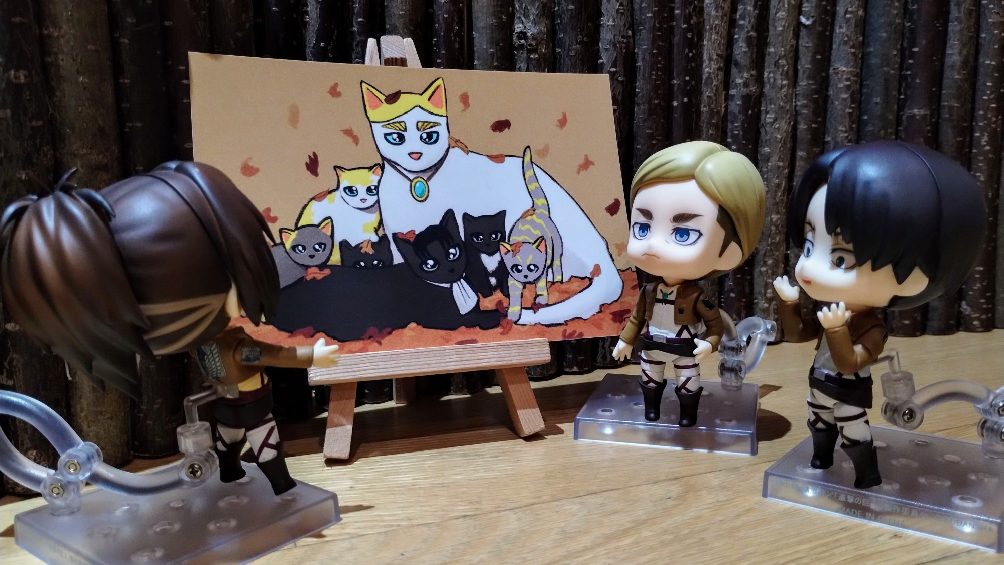 Nendoroids of Erwin Smith, Levi and Hange Zoe stand around a painting of a family of cats. Hange is turned towards the painting and is in a wide-legged, arms-outstretched posture indicating their surprise.