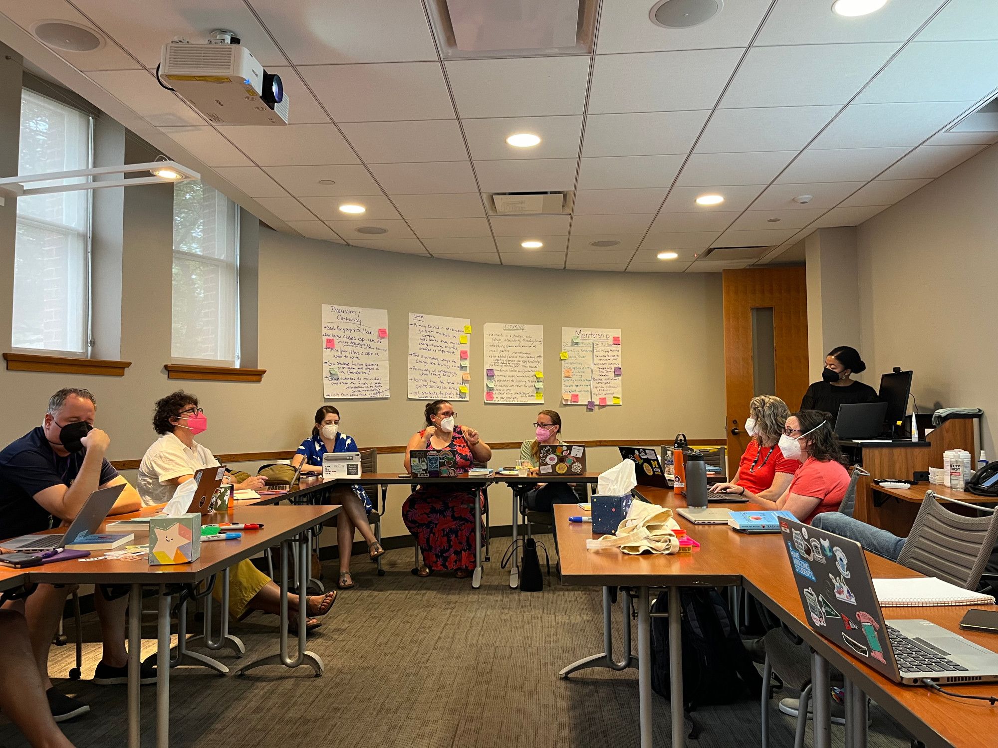 A photograph of the seminar room at The Bright Institute. Tables are arranged in a large rectangle around which the fellows are sitting. You can see us in conversation with Dr Tracie Addy, who stands at the computer desk on the right side of the room. Everyone is masked. On the far wall are posters we made about student engagement on Wednesday.