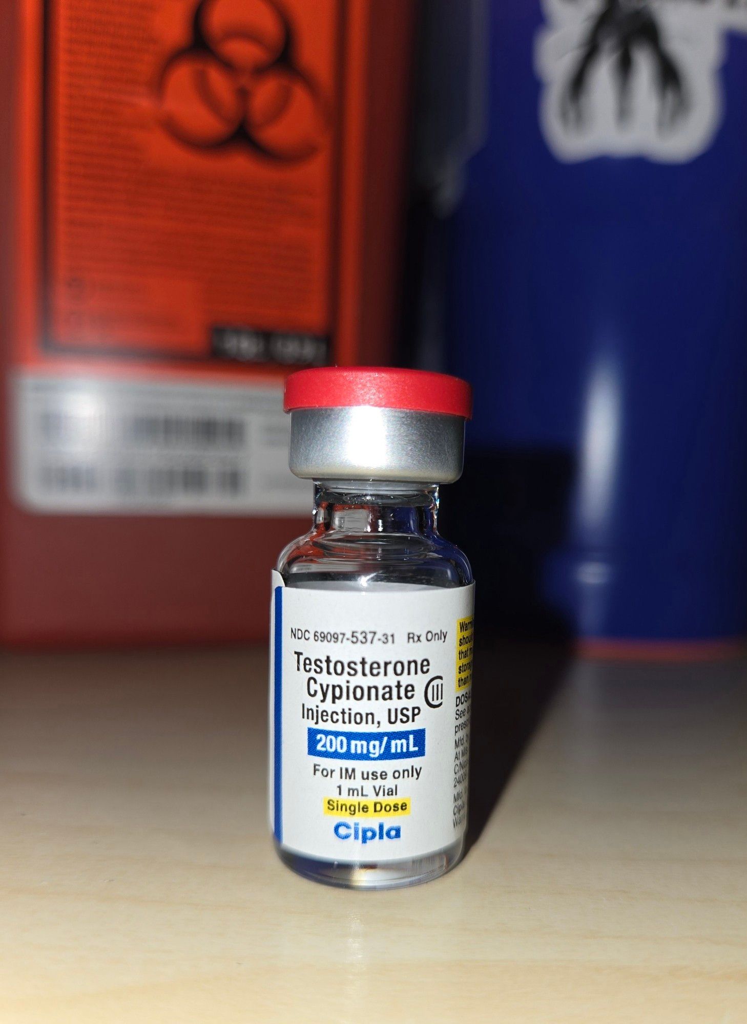 Little glass vial with a red safety cap and white label reading: testosterone cypionate, injection, USP, 200 mg/ml, for IM USE only. 1 mL Vial. Single dose. Cipla. (At the top is Rx only and a product code). Behind it is a blurry red sharps container with a biohazard symbol and a blurry blue-violet water cup with a Mass Effect Reaper sticker