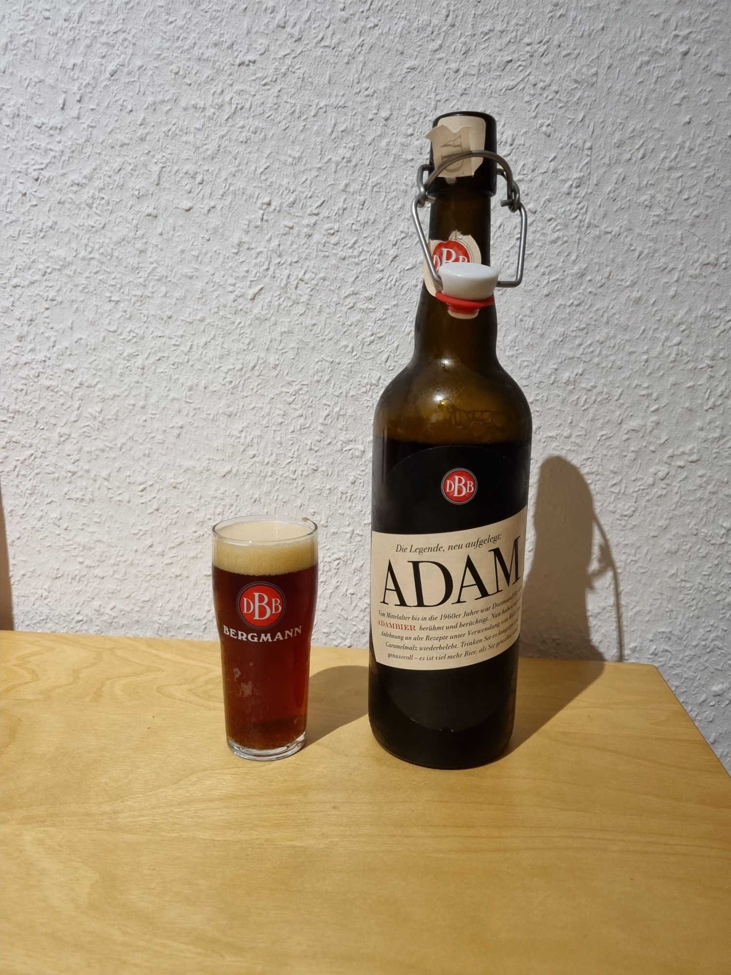 Ein geöffnete Bügelflasche Adam-Bier  und ein Stößchen (gefülltes 0,1 Liter Glas) der dortmunder Bergmann Brauerei auf Holztisch vor weißer Raufasertapete