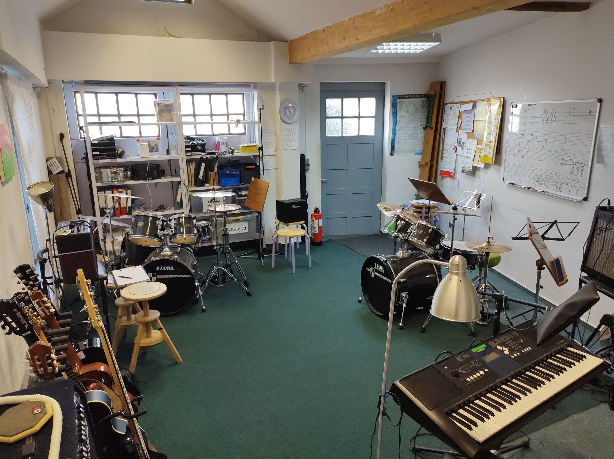 Ein Garagenraum mit drei Drumsets, Keyboard, Mikros,  jeder Menge Gitarren, E-Gitarren, E-Bässen, einen Regal voller Equipment und Unterrichtsunterlagen und diversen Infos an Pinwand, Whiteboard und auf diversen Postern. Alles ist aufgeräumt, Notenständer stehen vor den Instrumenten und zwei Mikros sind aufgebaut. Viele Verstärker und ein Mischpult sind angeschlossen. Alles ist quasi im Kreis angeordnet, die Mitte vom Raum isr frei.