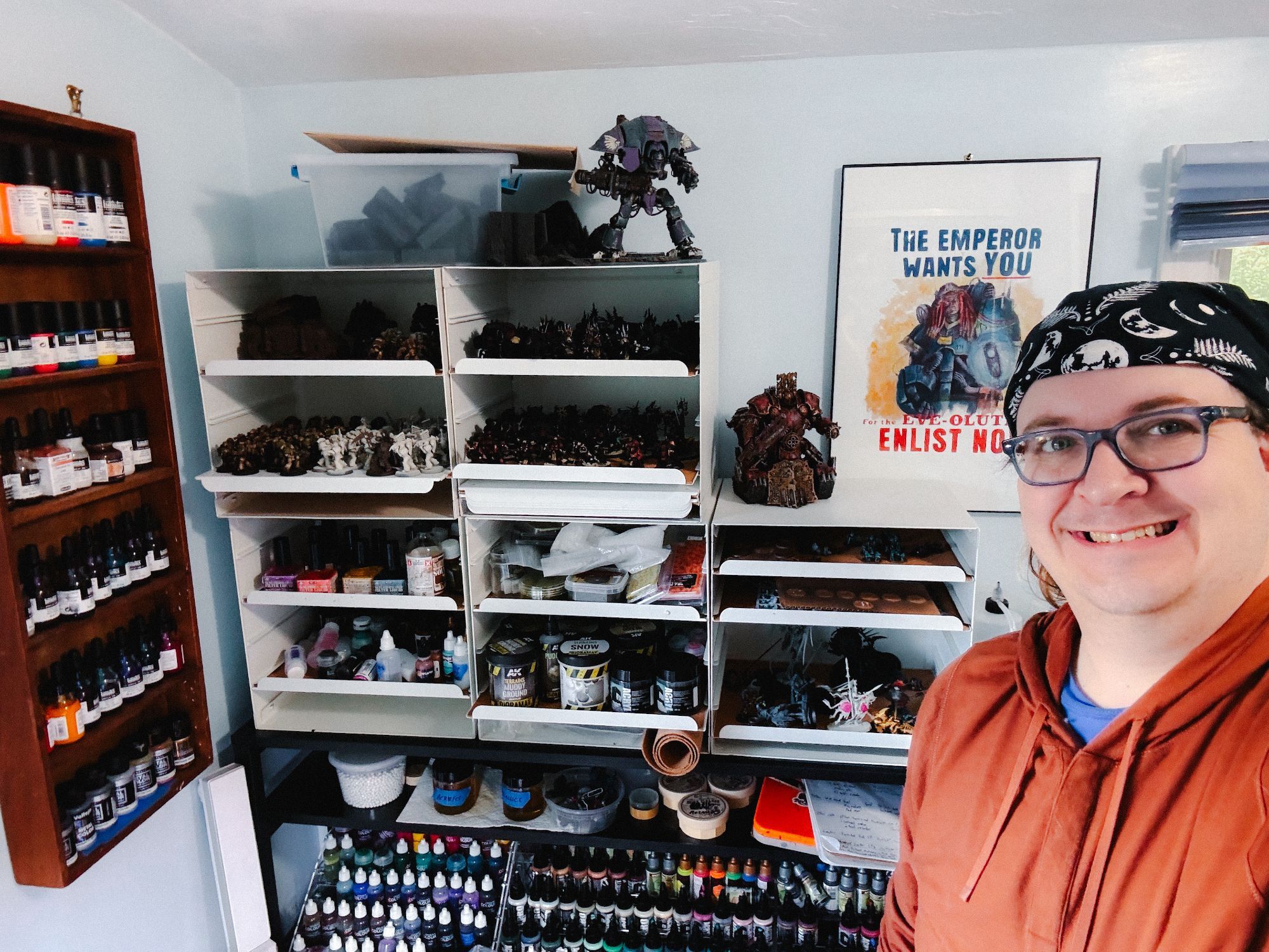 Selfie, with me standing in front of my miniatures painting hobby area. Behind me are an embarrassing number of colorful small bottles of paint (like 150 maybe) in nail polish racks and a wooden wall rack, plus five stacked Ikea letter cabinets full of more paints and models. On the wall behind me is a original poster showing a female space marine in blue power armor, with the text "The Emperor Wants You for the Eve-olution. Enlist now."