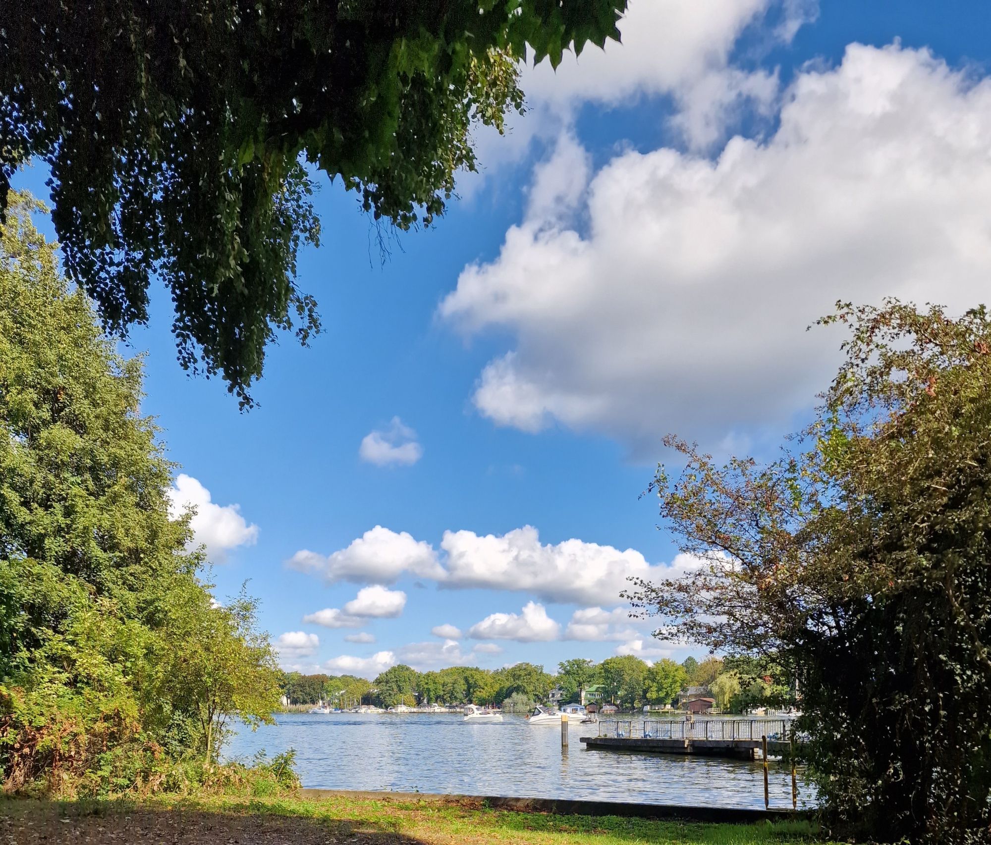 Dahme (Fluss) im Süden Berlins. Aus dem Dunkel unter Bäumen heraus fotographierter Flussblick mit beiden Ufern. Viel Grün, Schiffe und Boote in der Ferne erkennbar. Darüber ein intensivblauer Himmel mit dicken, wattigen Wolken.