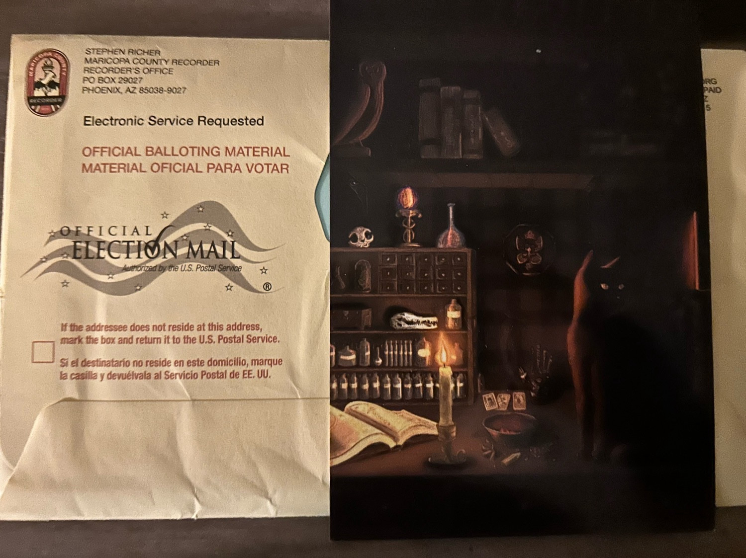 A photo of a mail in ballot, unopened, from maricopa county. There's a cute postcard of a black cat in a spooky house covering the right half, where all the personal info is