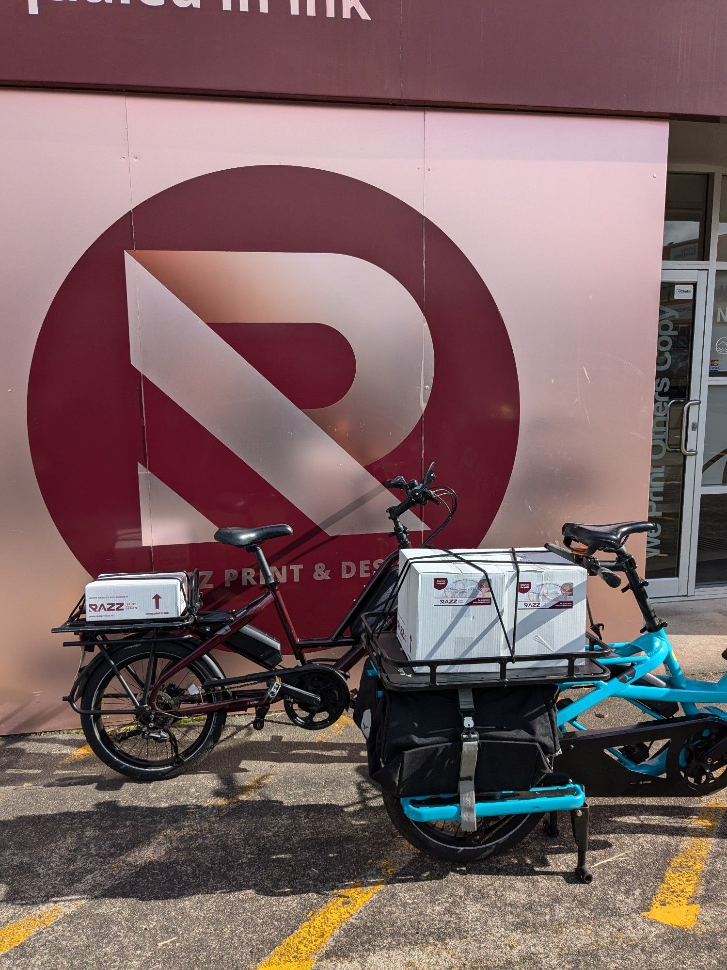 Two E-bikes parked outside a store loaded with boxes of printing.