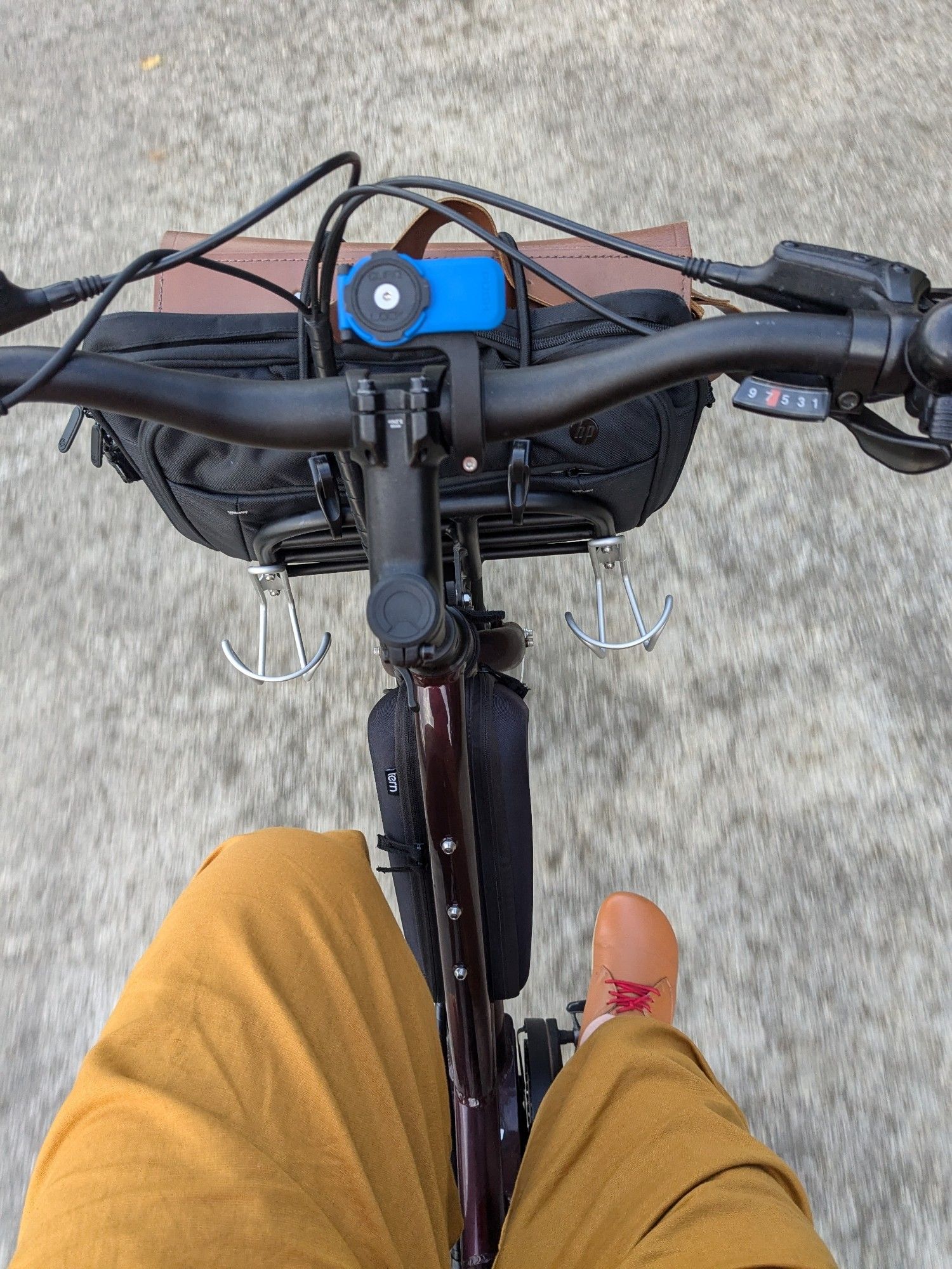 Weird angle picture of a bike showing handlebars, laptop bag and satchel strapped to the front tray, yellow pants and brown shoes of the rider.