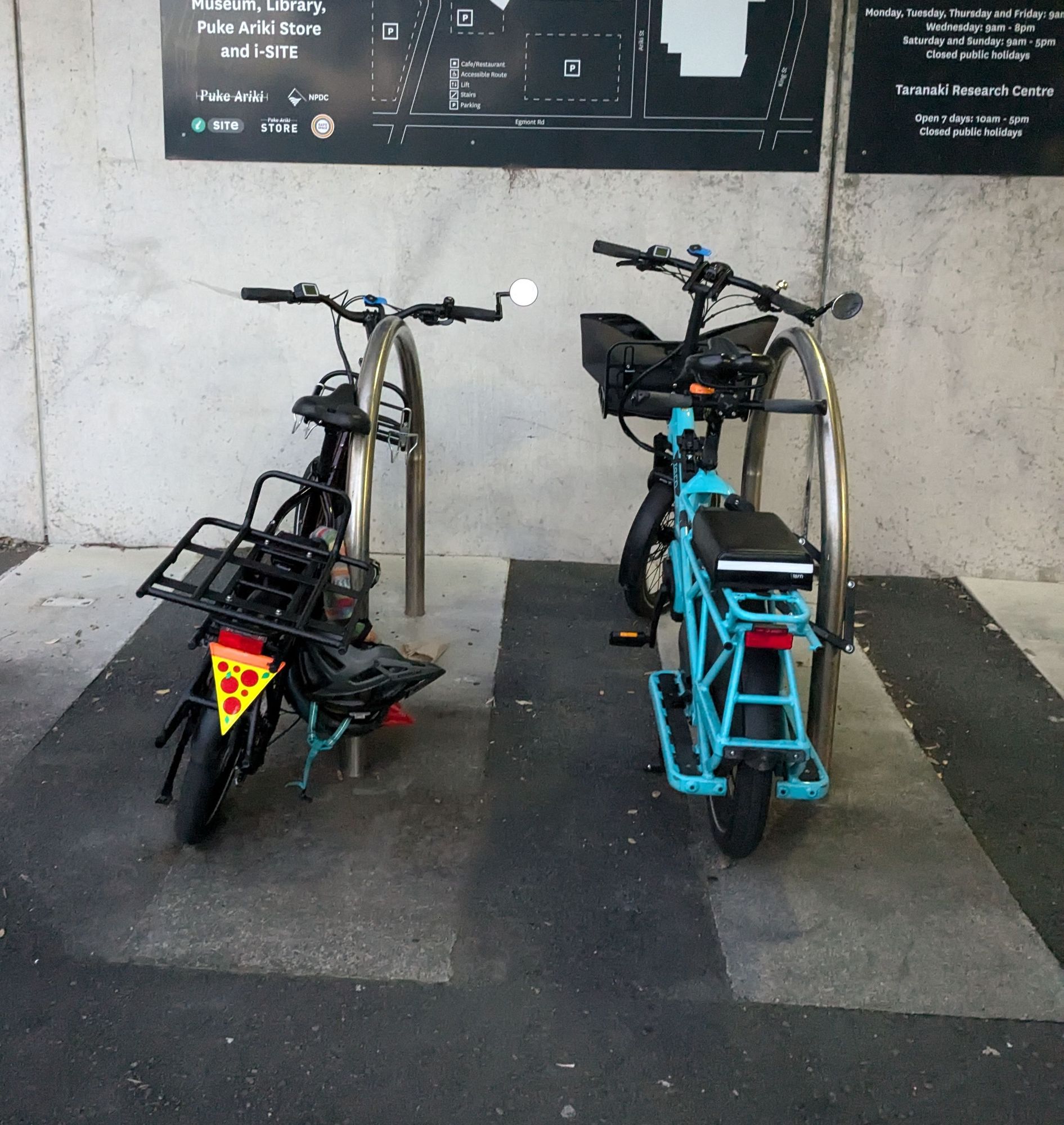 Two bikes parked at bike racks. #BoysenberryTheBike is a Tern Quick Haul with rack and a safety flag on the back shaped like a piece of pizza. #BlueberryTheBike is a blue Tern GSD with a child seat on the back.