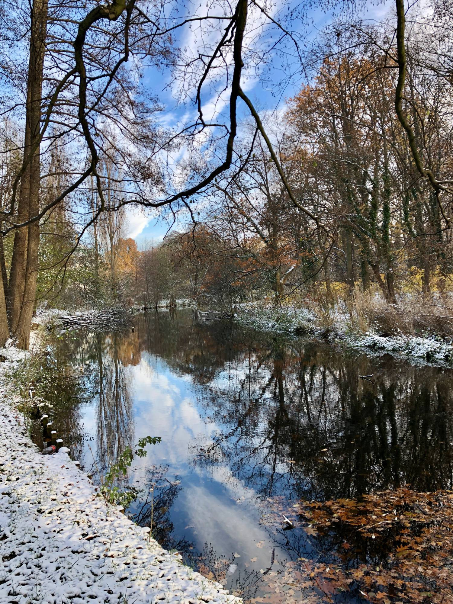 Blick auf einen Wasserlauf, an dessen Ufern Bäume und Büsche stehen. Bauer Himmel. Es liegt Schnee und alles spiegelt sich im Wasser.