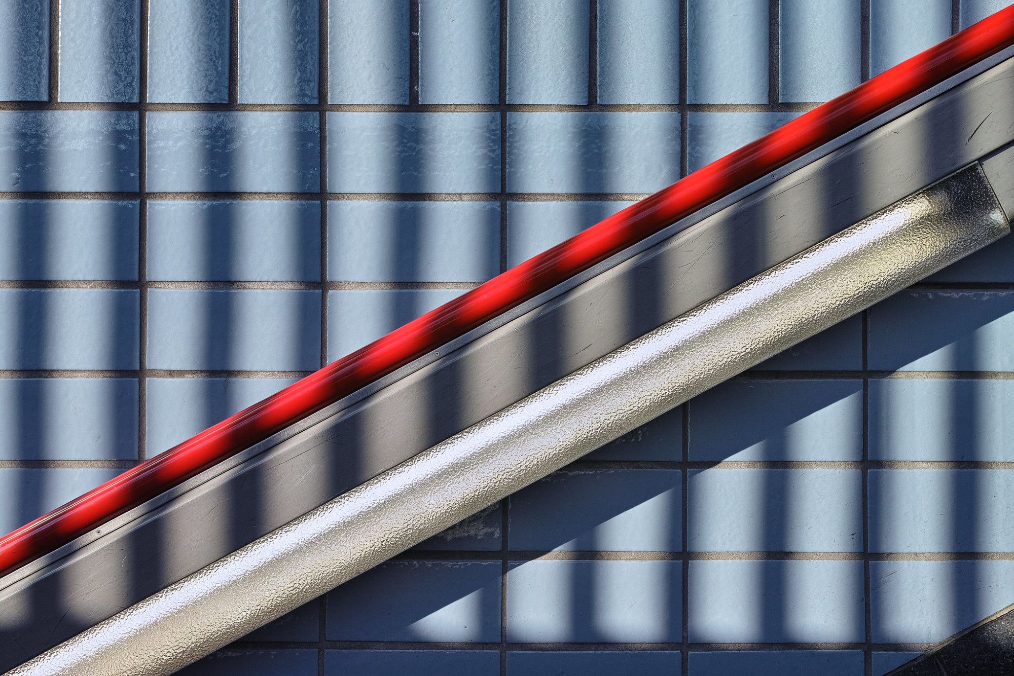 Wand mit rechtechigen blauen Kacheln. Darauf dere Schatten eines  Geländers mit senkrechten Streben. Davor ein Handlauf mit roter Leiste oben und darunter eine Leuchtstoffröhre.