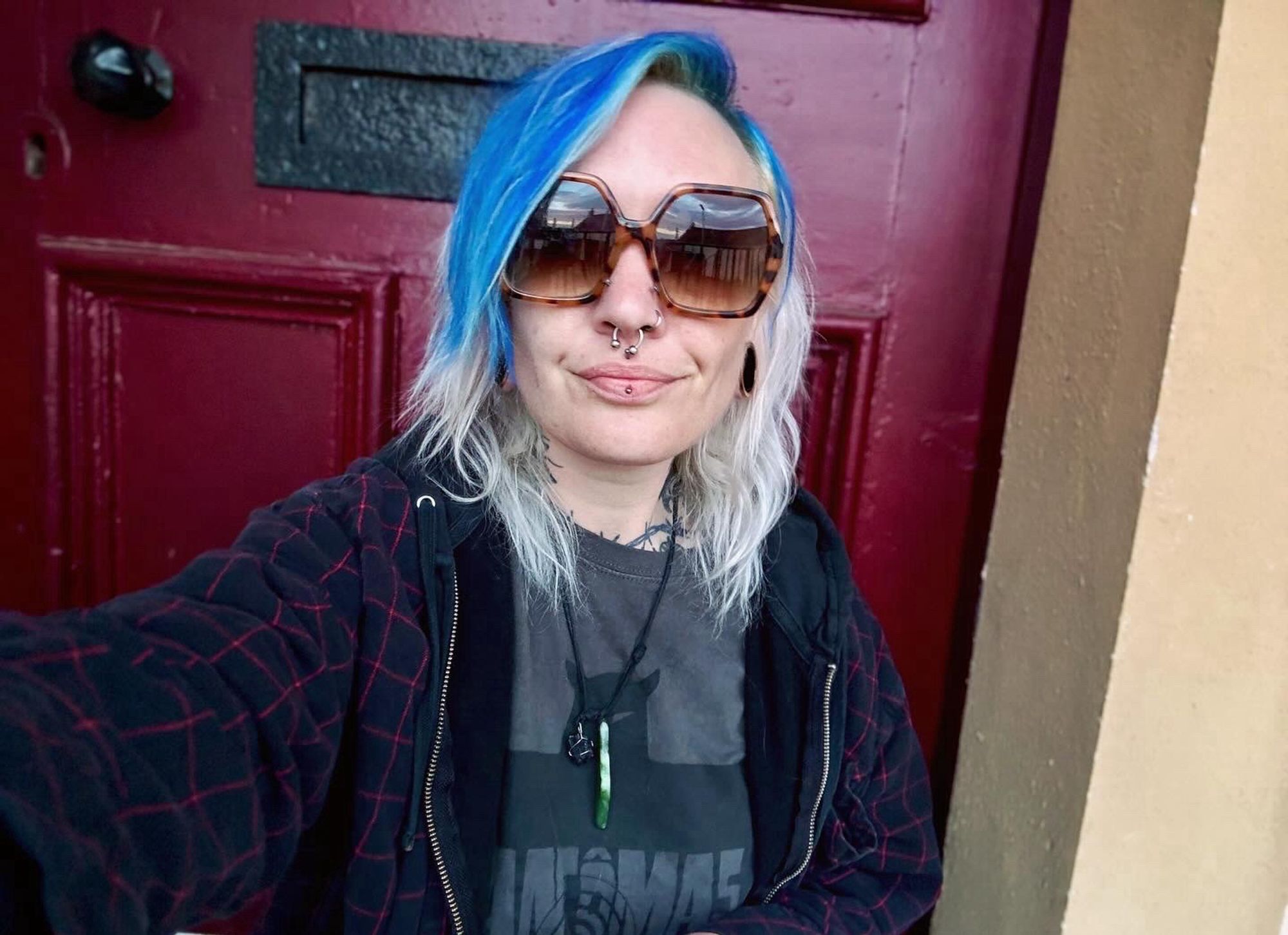 Selfie of Katie sat in front of a maroon coloured door. Her hair is bright blue at the front and white blonde at the back.