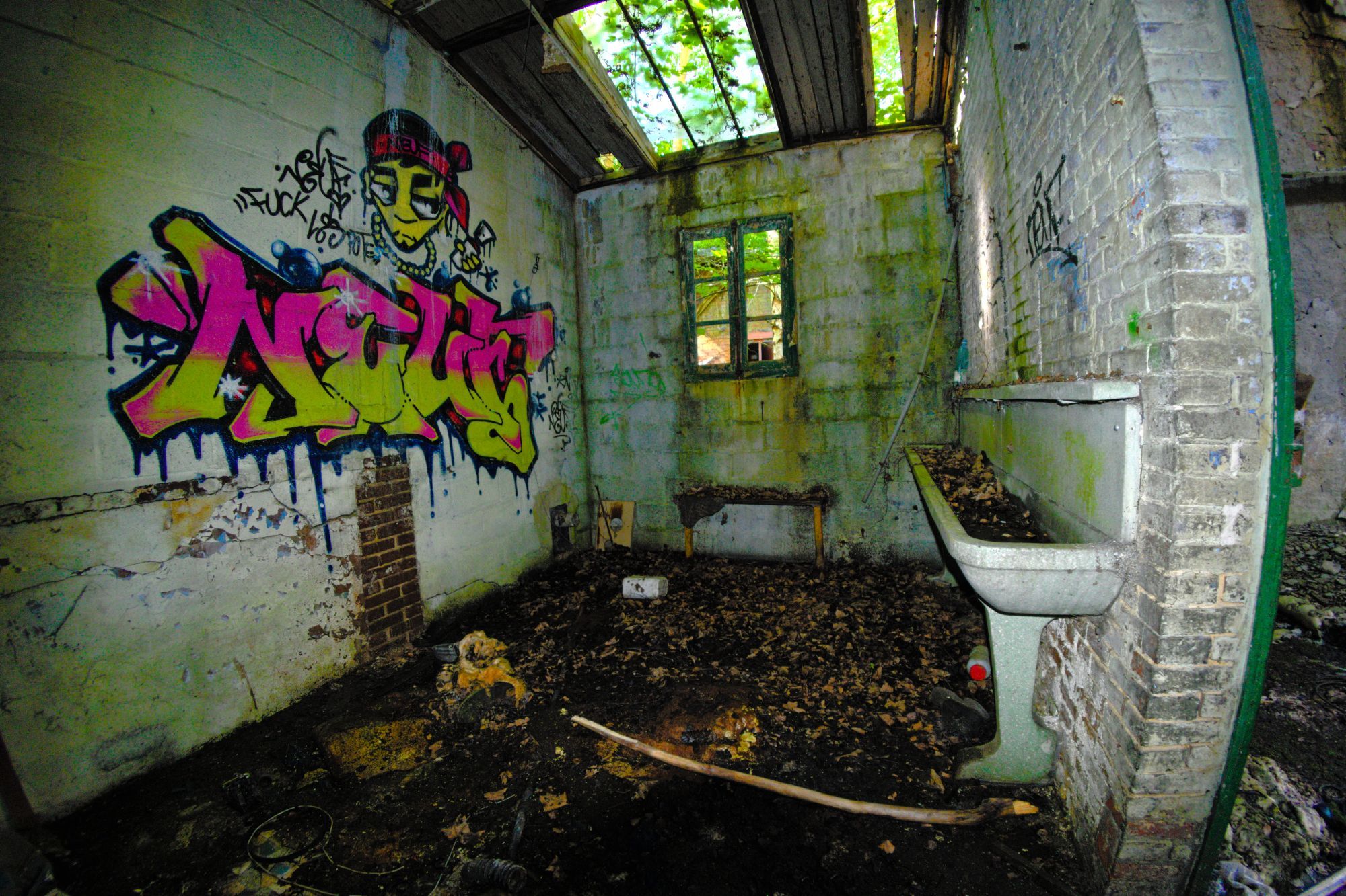 Une salle de sanitaires dans une usine en ruine. Sur la droite, contre un mur, un lavabo collectif. Sur la gauche, sur le mur opposé, un graphe. Le plafond est défoncé. Le sol est couvert de feuilles mortes et de déchets.