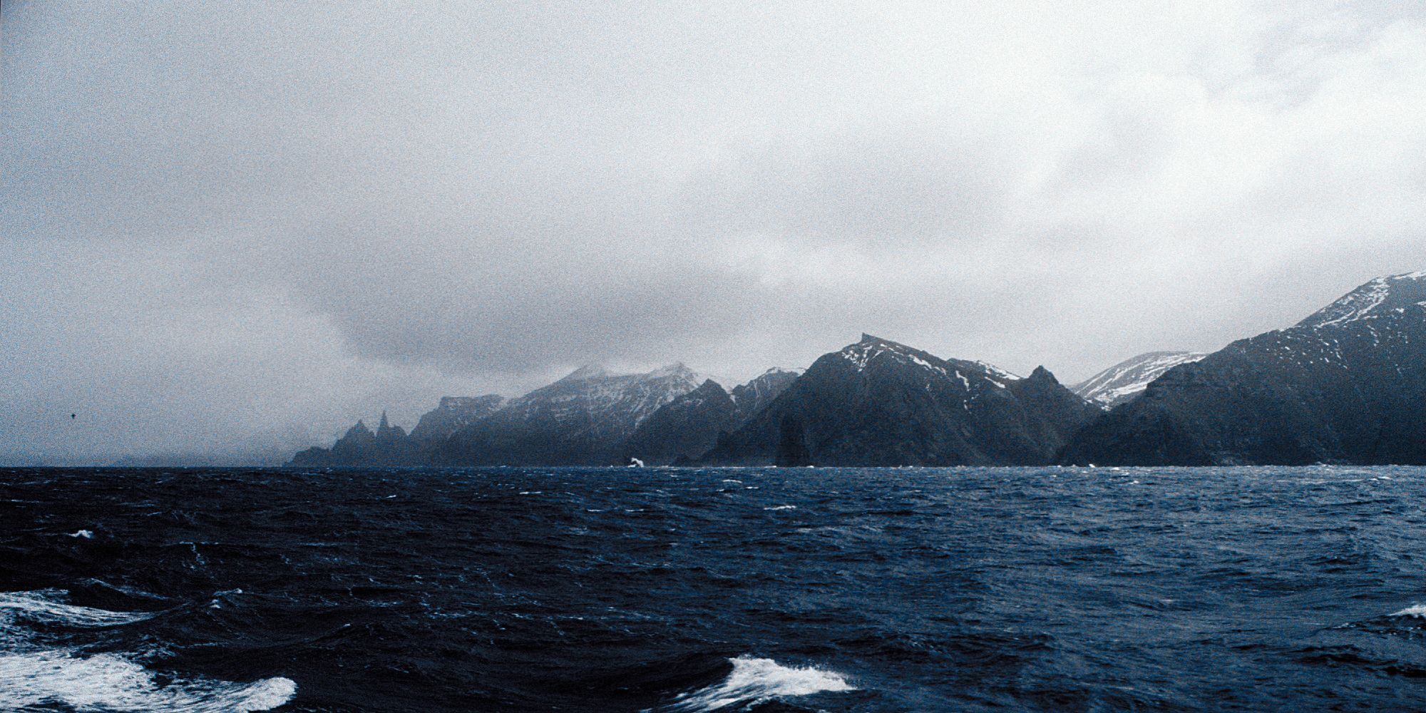 Des montagnes noires couvertes de quelques taches de neiges plongent vers une mer sombre et agitée. Le ciel est nuageux. La photo est prise de la mer.