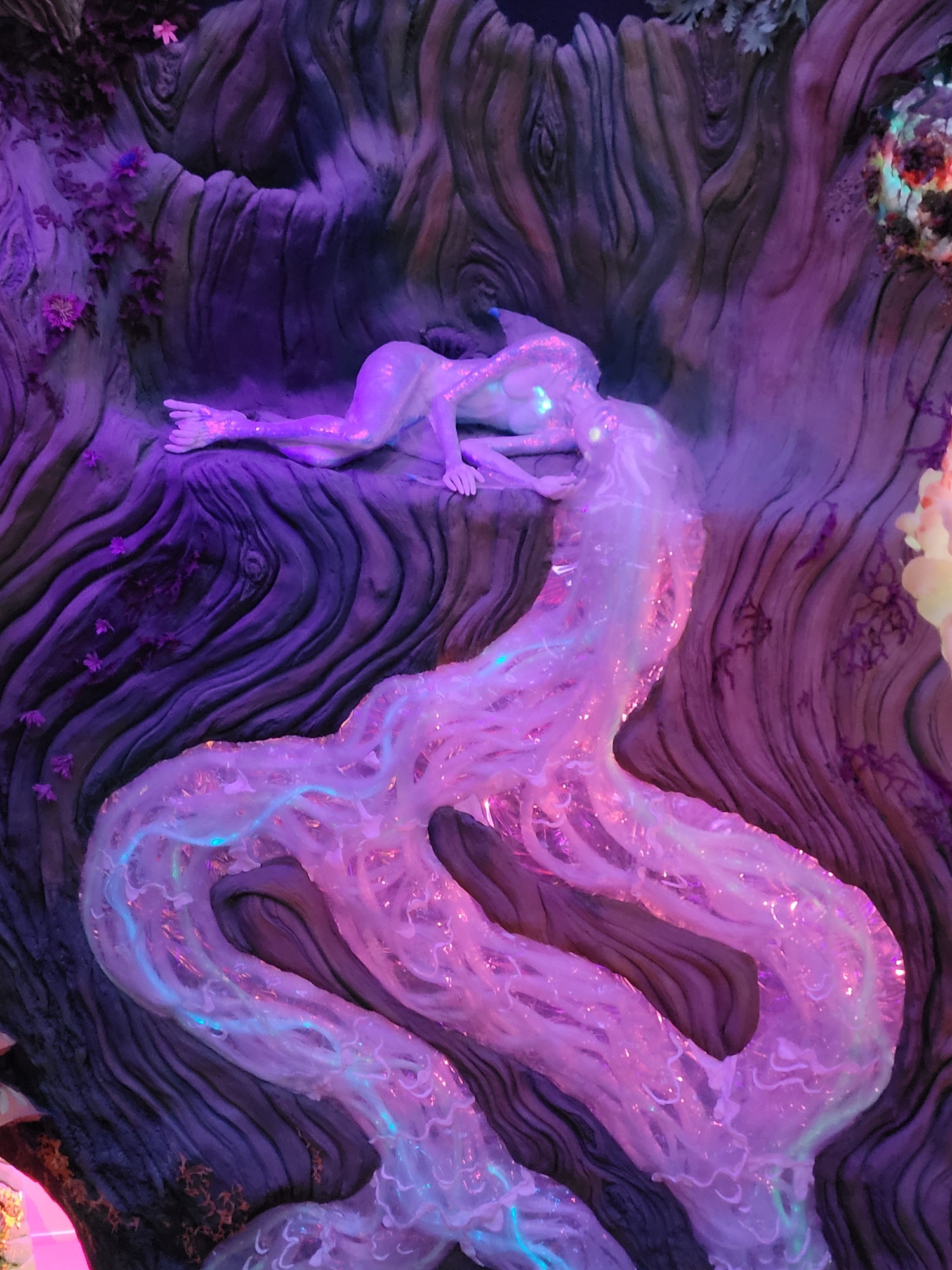 Translucently white feminine figure laying on her side on the nook in what might be a tree. Her head/hair flows into branching streams down the trunk