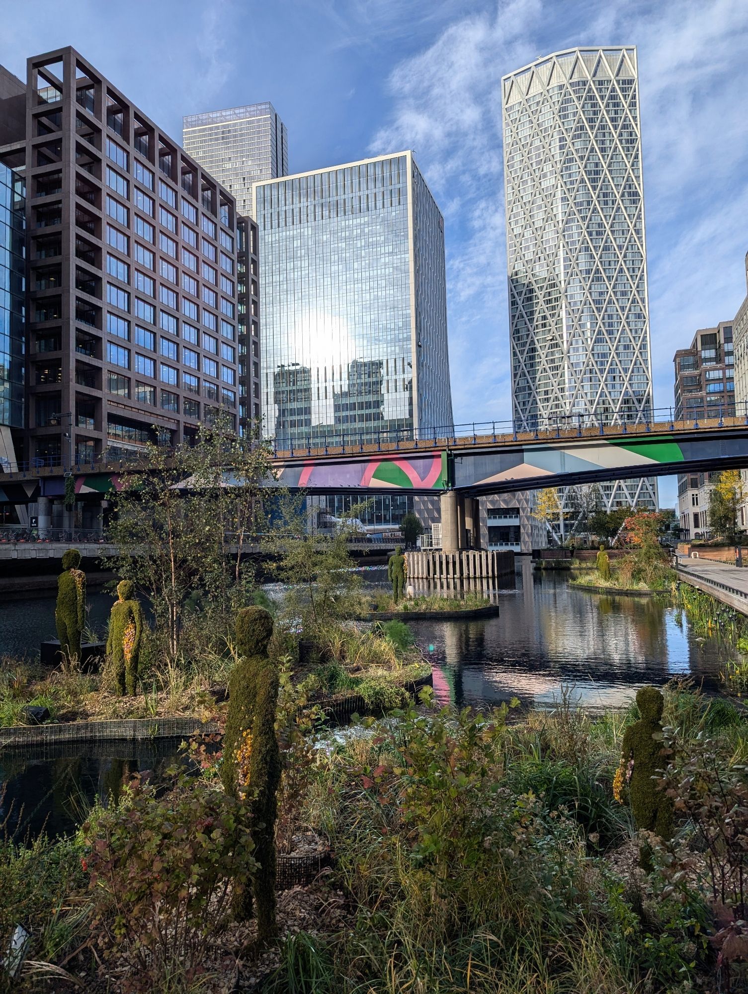 A photo of Canary Wharf in London, UK. Towers and a bridge surround a watery wharf. In the foreground, human-shaped figures made of plants stand in patches of foliage.