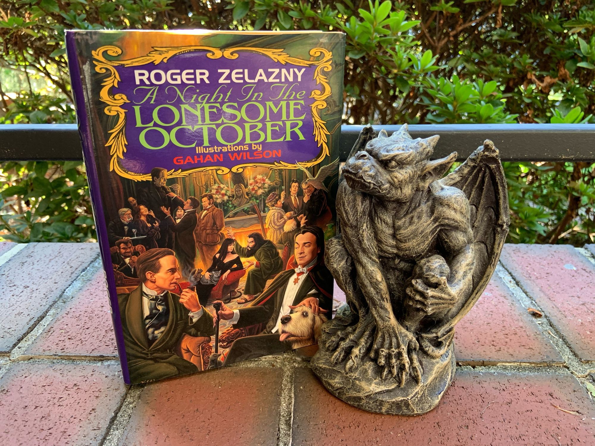 A picture of A NIGHT IN THE LONESOME OCTOBER (with cover art depicting a meeting of many familiar Gothic characters, including Sherlock Homes and Count Dracula in the forefront) sitting on a brick walk beside a stone gargoyle.