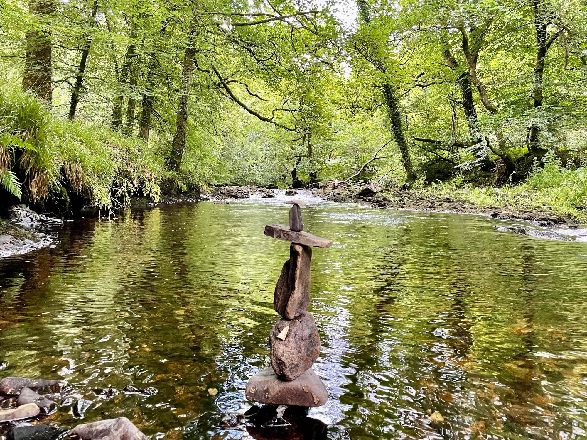 River Teign, Devon, England. At the fringes of a woodland river, stones have been carefully stacked to form the shape of a cross.