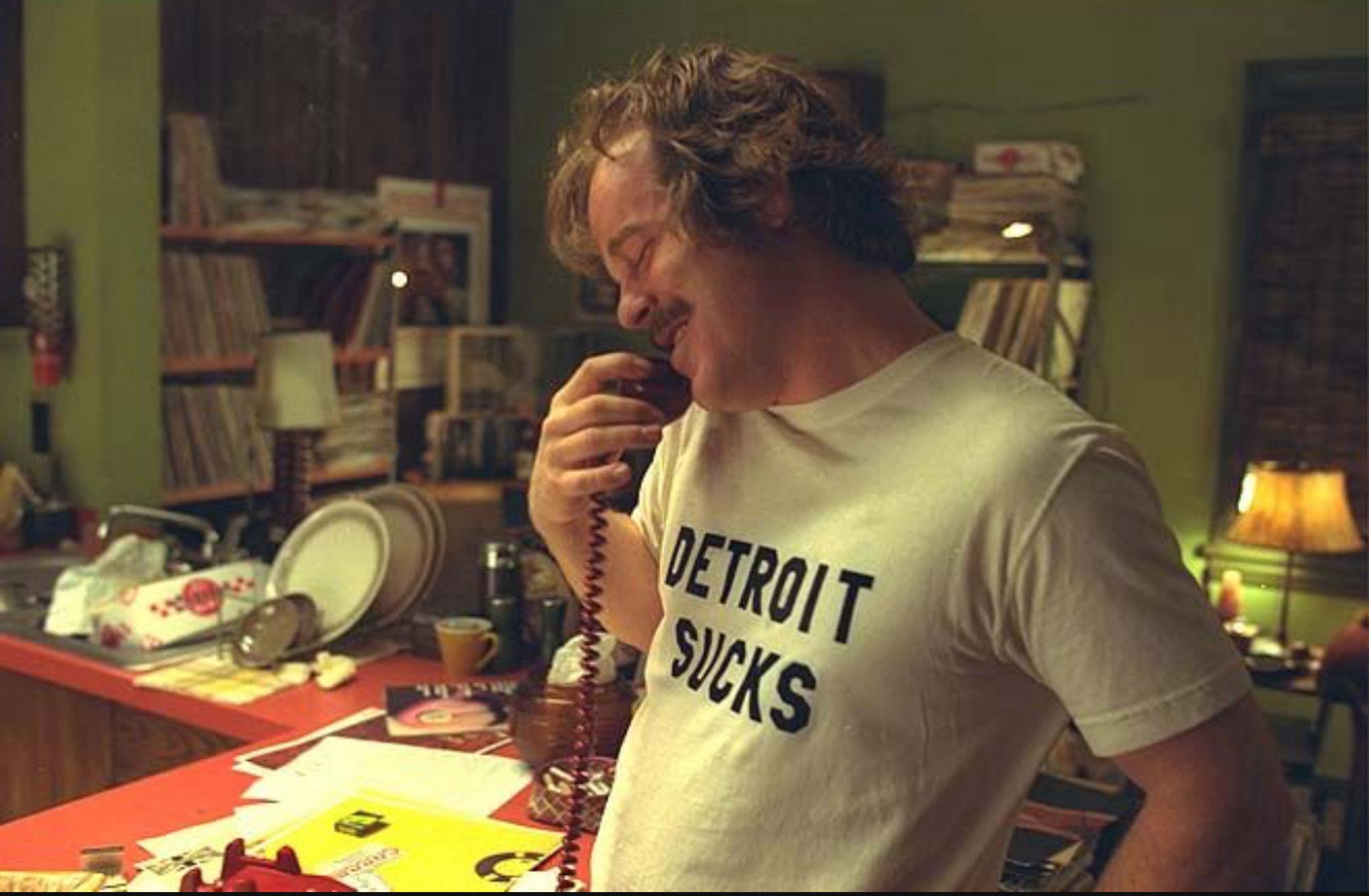 Philip Seymour Hoffman as Lester Bangs in Almost Famous. He’s talking on a telephone and wearing a white t-shirt that says, “Detroit Sucks”.