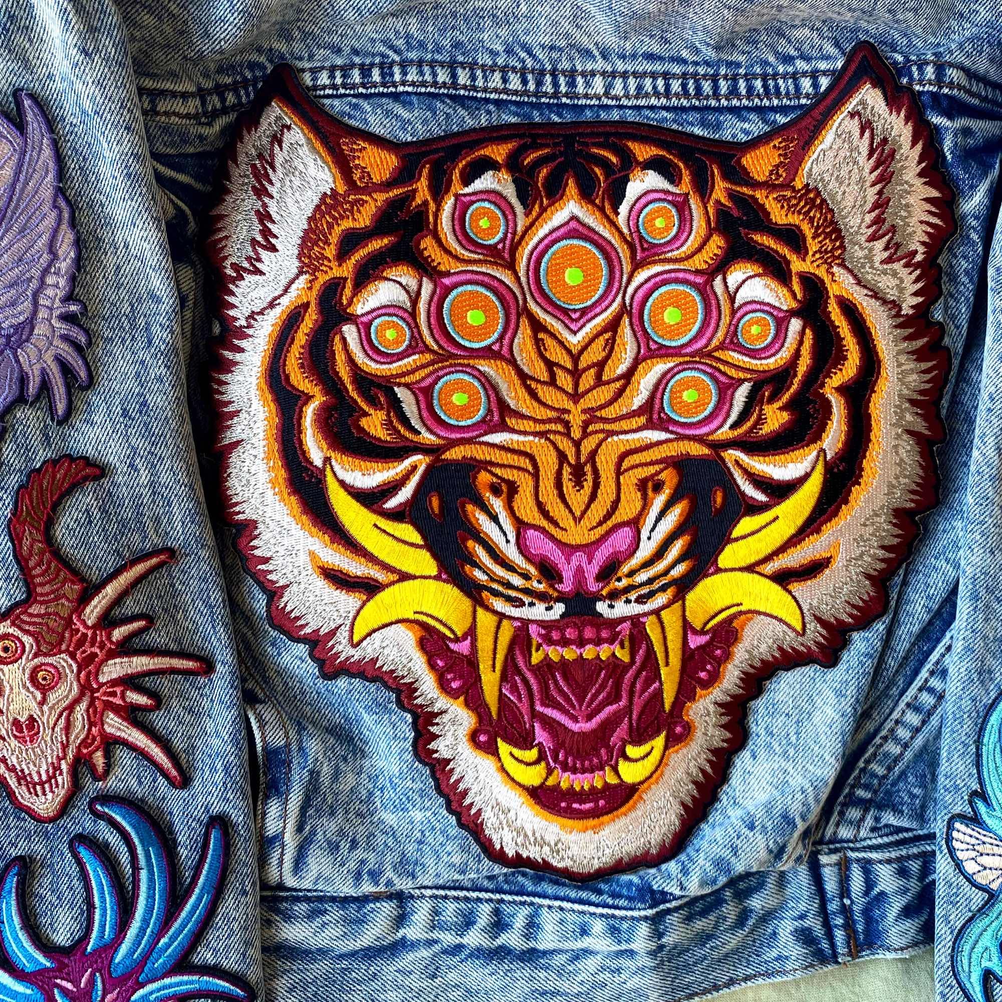 Photo of a denim jacket with colorful patches attached. Featured is a large patch depicting a tiger head with many eyes and large teeth