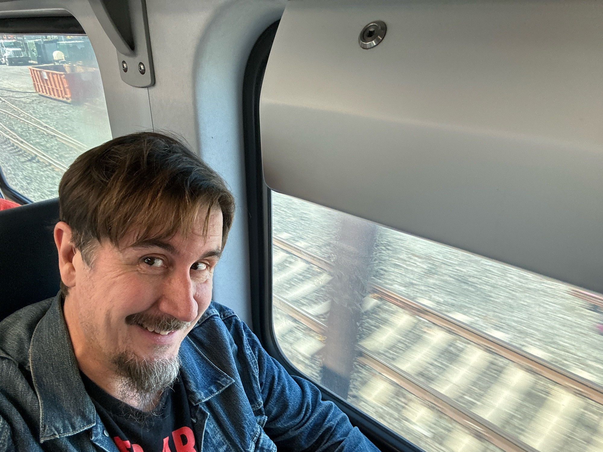 Picture of John Darnielle, a middle-aged guy with a goatee and the best hair of really anybody, on a NJ line train. He’s arena rock casual in a blue Levi’s jacket over an inter arma tee. Behind him, through the window, the tracks, a blur of motion. 