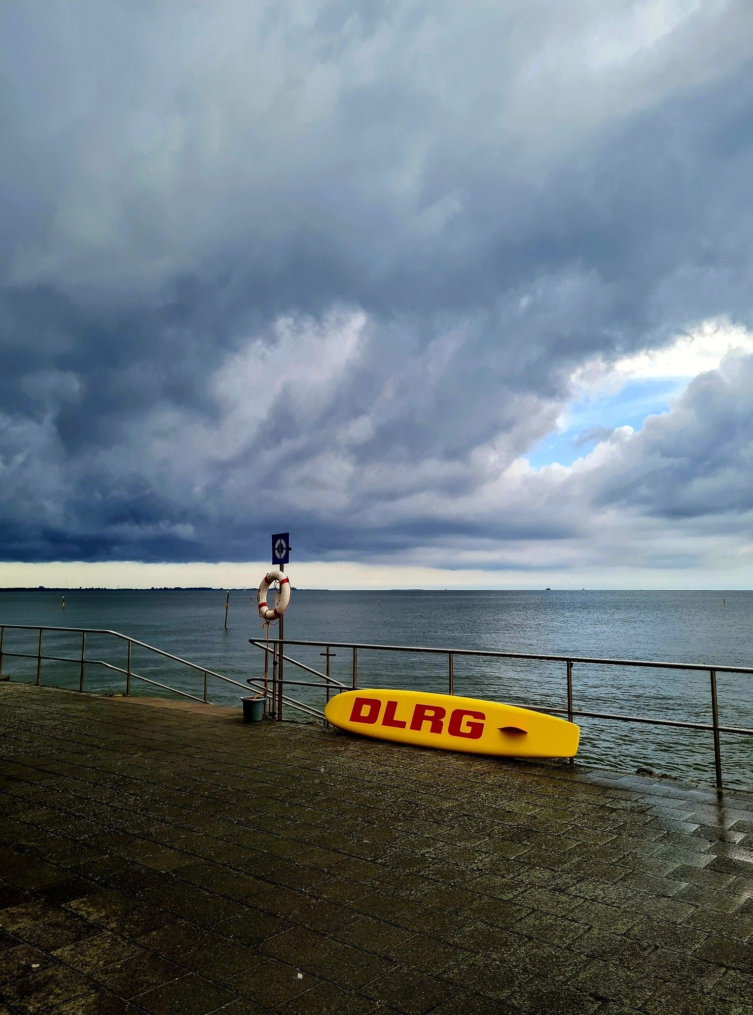 Farbfoto im Hochformat: Blick aufs Meer bei Regenwetter. Im Vordergrund grau gepflasterte Steine, dahinter ein Metallzaun. An einer Metallstange hängt ein weiß-roter Rettungsring, vor dem Zaun lehnt ein gelbes Rettungsbrett mit roten Buchstaben, darauf steht DLRG. Das Meer sieht dunkel aus. Mehr als die Hälfte des Bildes nimmt der Himmel ein, der mit dunklen Gewitterwolken gefüllt ist. Am rechten Bildrand befindet sich in den dunklen Wolken ein Loch, durch das ein kleines Stück blauer Himmel
sichtbar wird.