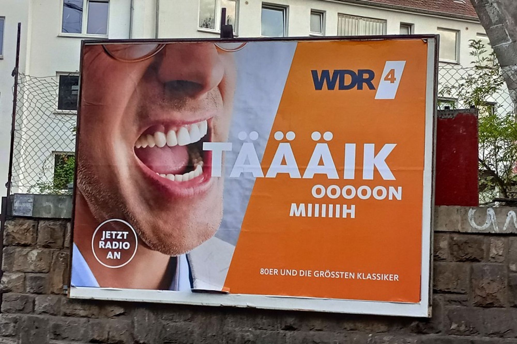 Plakatwand mit einem Werbeplakat des Radiosenders WDR4. Darauf abgebildet ist ein (oben und seitlich angeschnittenes) Gesicht eines offenbar singenden Mannes, daneben steht auf orangefarbigem Hintergrund der Text "TÄÄÄIK OOOOON MIIIIIH".