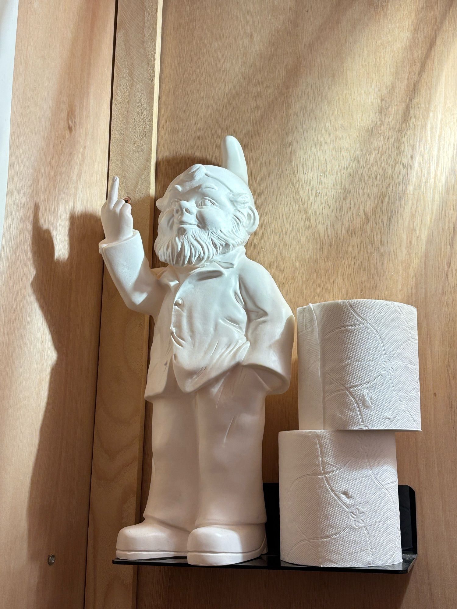 A white plaster bearded gnome with his middle finger sticking up stands on a bathroom shelf beside two spare rolls of toilet paper.