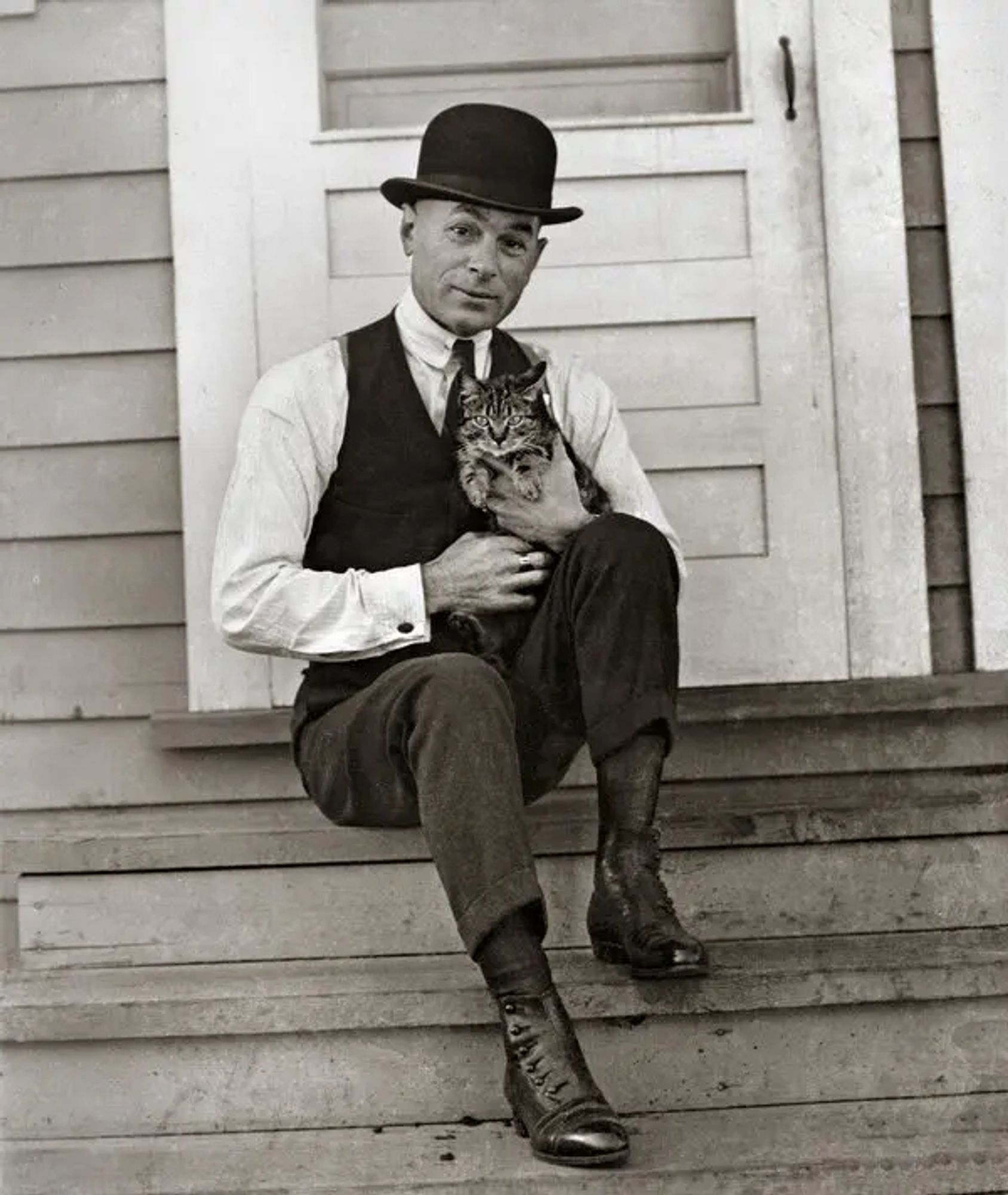 A man (a great cartoonist) sitting on some steps holding a cat. He's wearing a vest and tie and a derby hat.