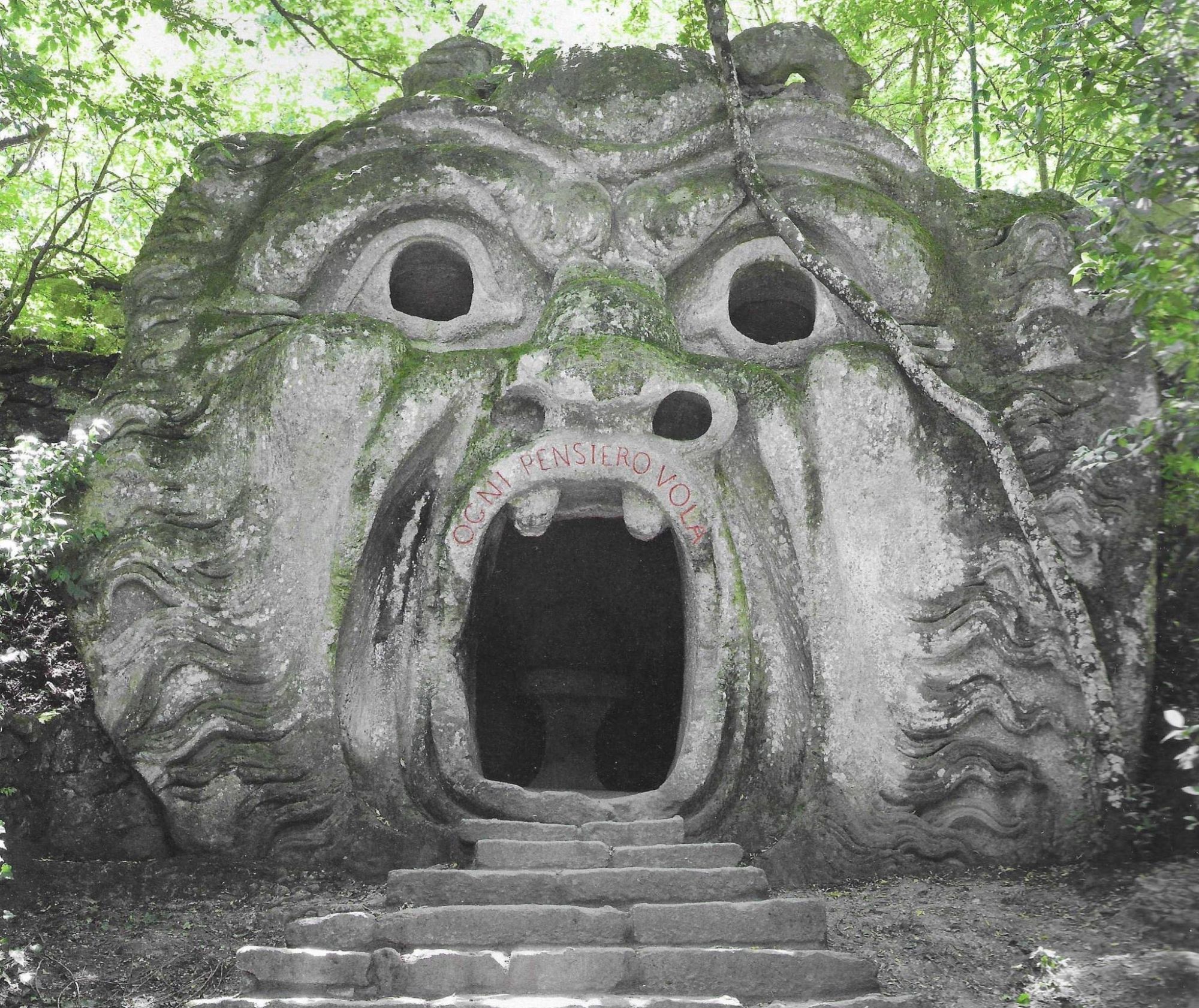 A stone Gateway sculpted into an enormous face are people enter up a flight of stairs into escaping mouth under a couple of large incisors pointing down at them on the lip of the monster and the words in Italian every thought flies