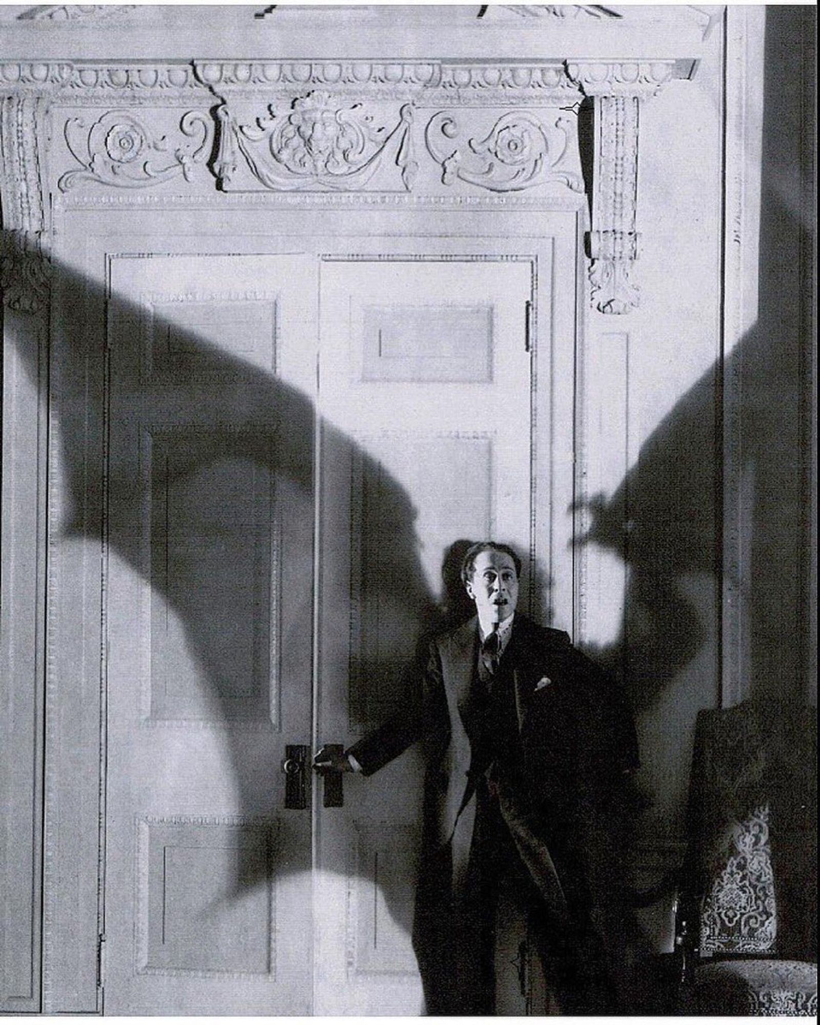 Film still (or promotional photo) Man with astonished look stopped with his hand on the knob of a double door staring at a figure with enormous bat wings whose shadow covers him.