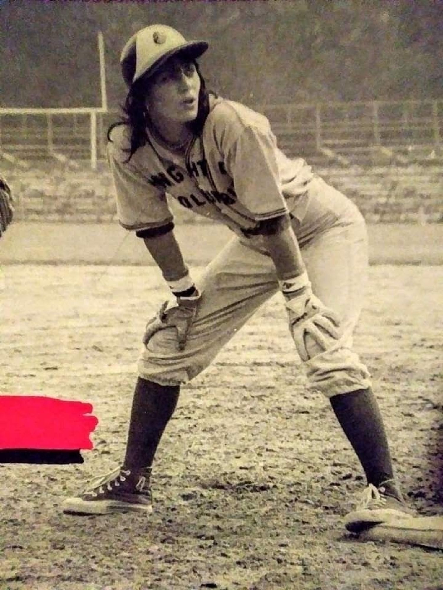 Black and white photograph of Joan Jett playing baseball is on base bent over hands on knees watching to see what the pictures doing she's wearing a baseball uniform with Knights of Columbus across the chest