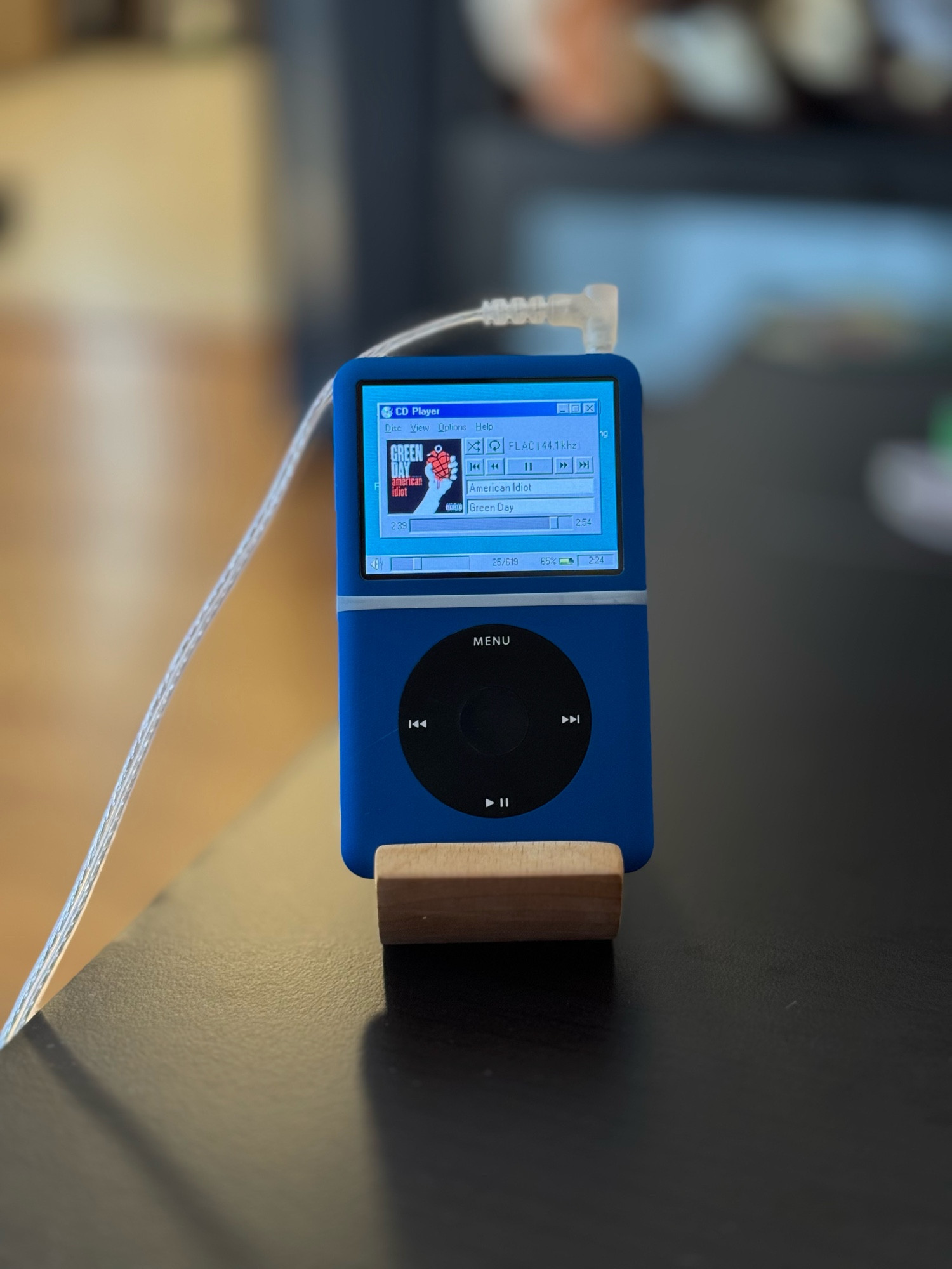 blue ipod classic with black clickwheel sitting on a stand on a table, displaying a rockbox now playing screen that looks like the windows 95 cd player app playing green day’s american idiot