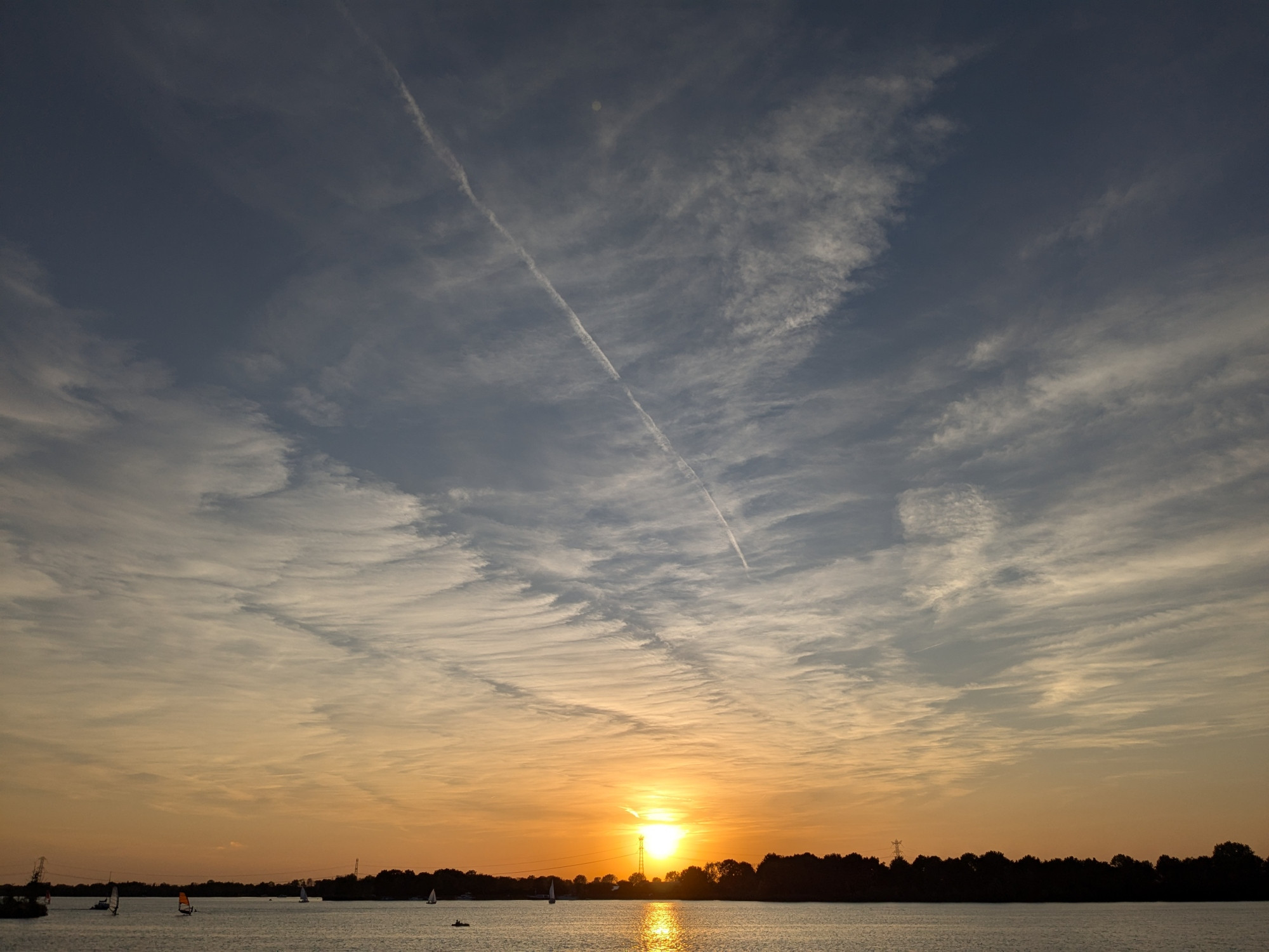 The sun is setting behind a lake with some boats and surfers. It turns the horzion yellowish and above the sky is blue with hazy, widened striped wispy clouds. At the horizon is a treeline.