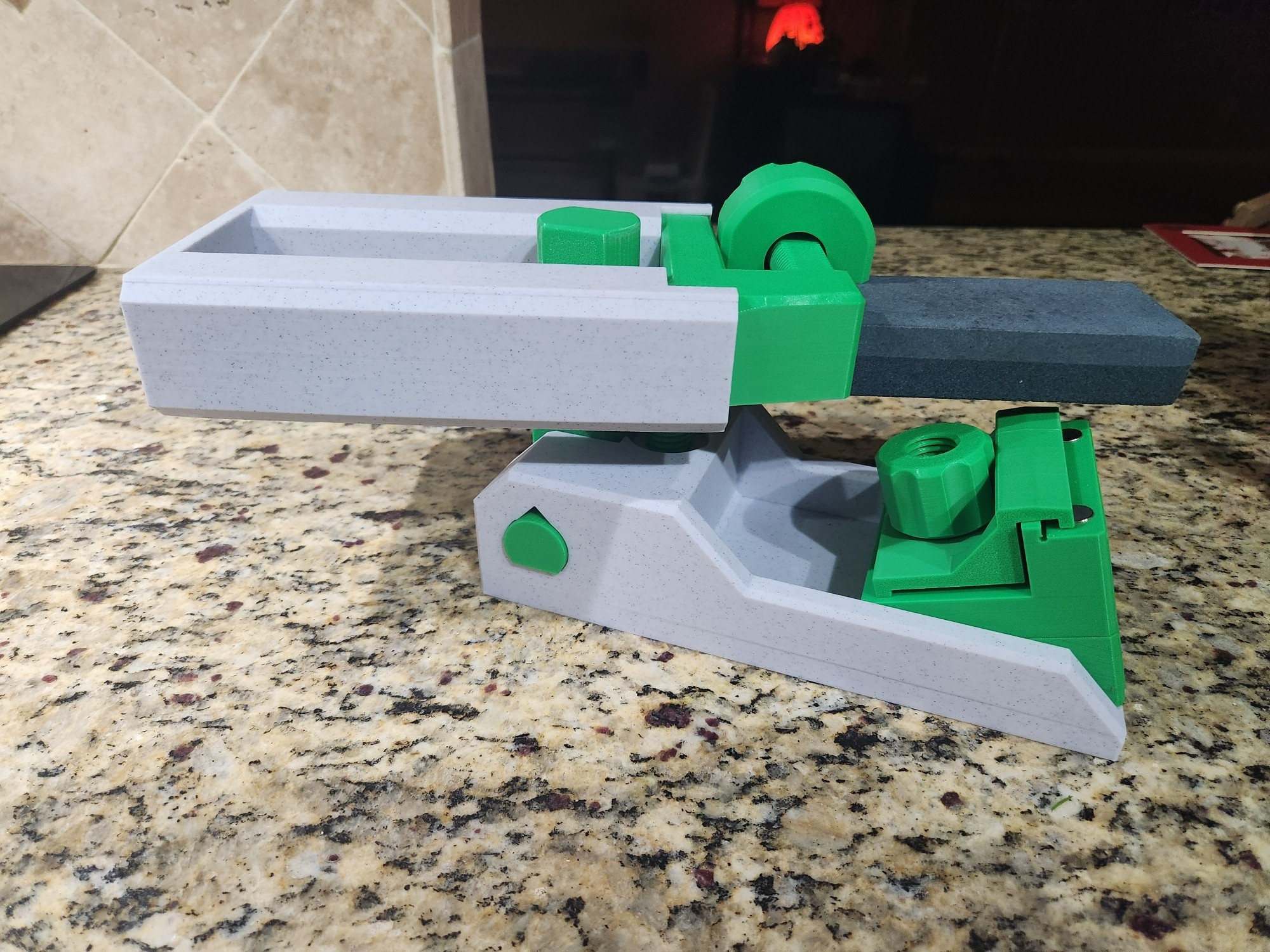 Stone grey and green swing arm knife sharpener. A whetstone is clamped on a guide rail swing arm to drag it over a knife clamp at 20 degrees. The bottom angle block can be replaced for a diff angle. The knife clamp has two neodymium magnets for easy set up and small knives.