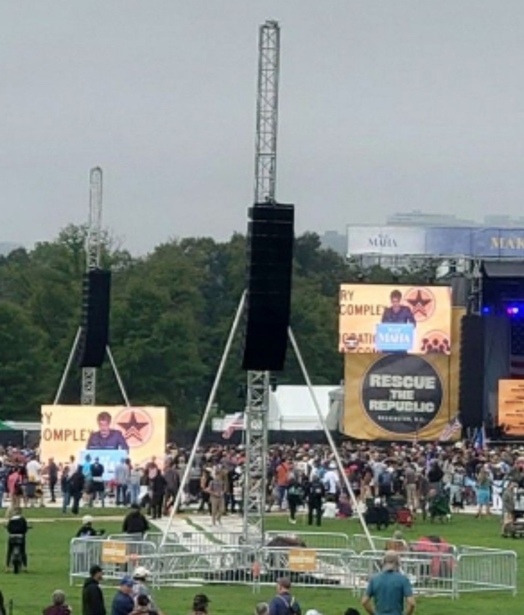 A tower of speakers meant to be in a music festival crowd stands alone and is pointed at no one. The crowd peters out around the speaker tower closer to stage.